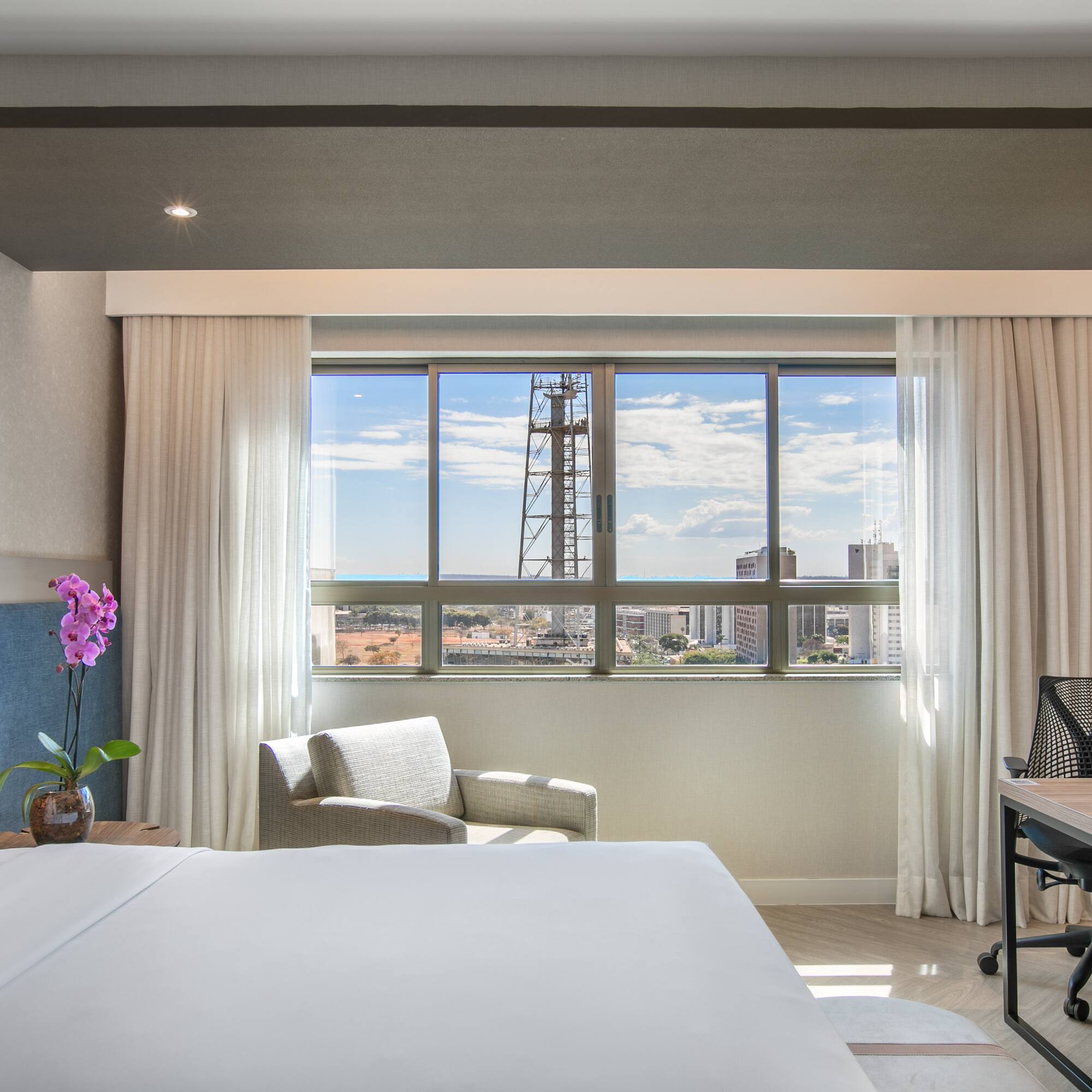 Modern hotel room with city view, white bed, armchair, and work desk.