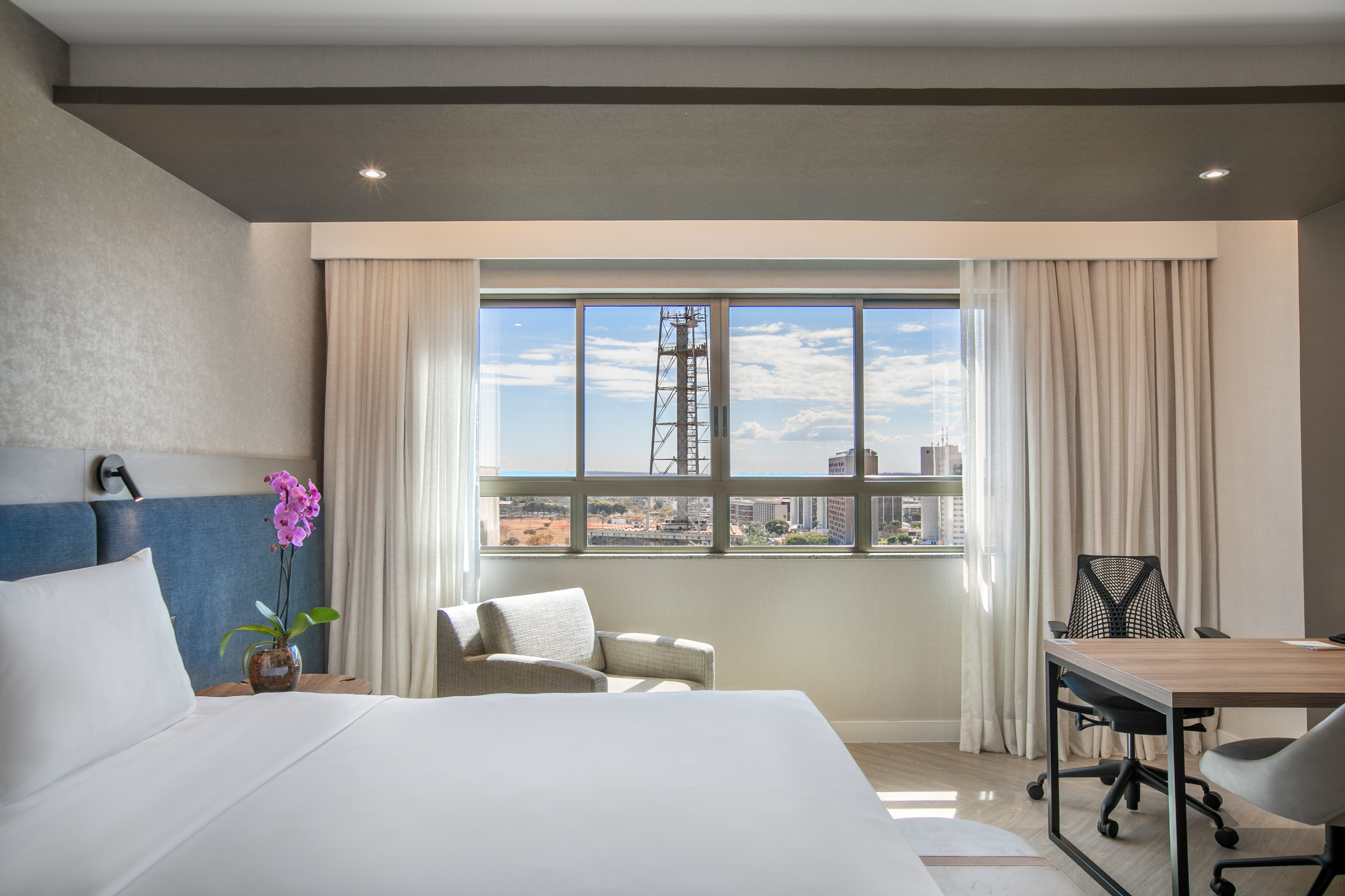 Modern hotel room with city view, white bed, armchair, and desk.