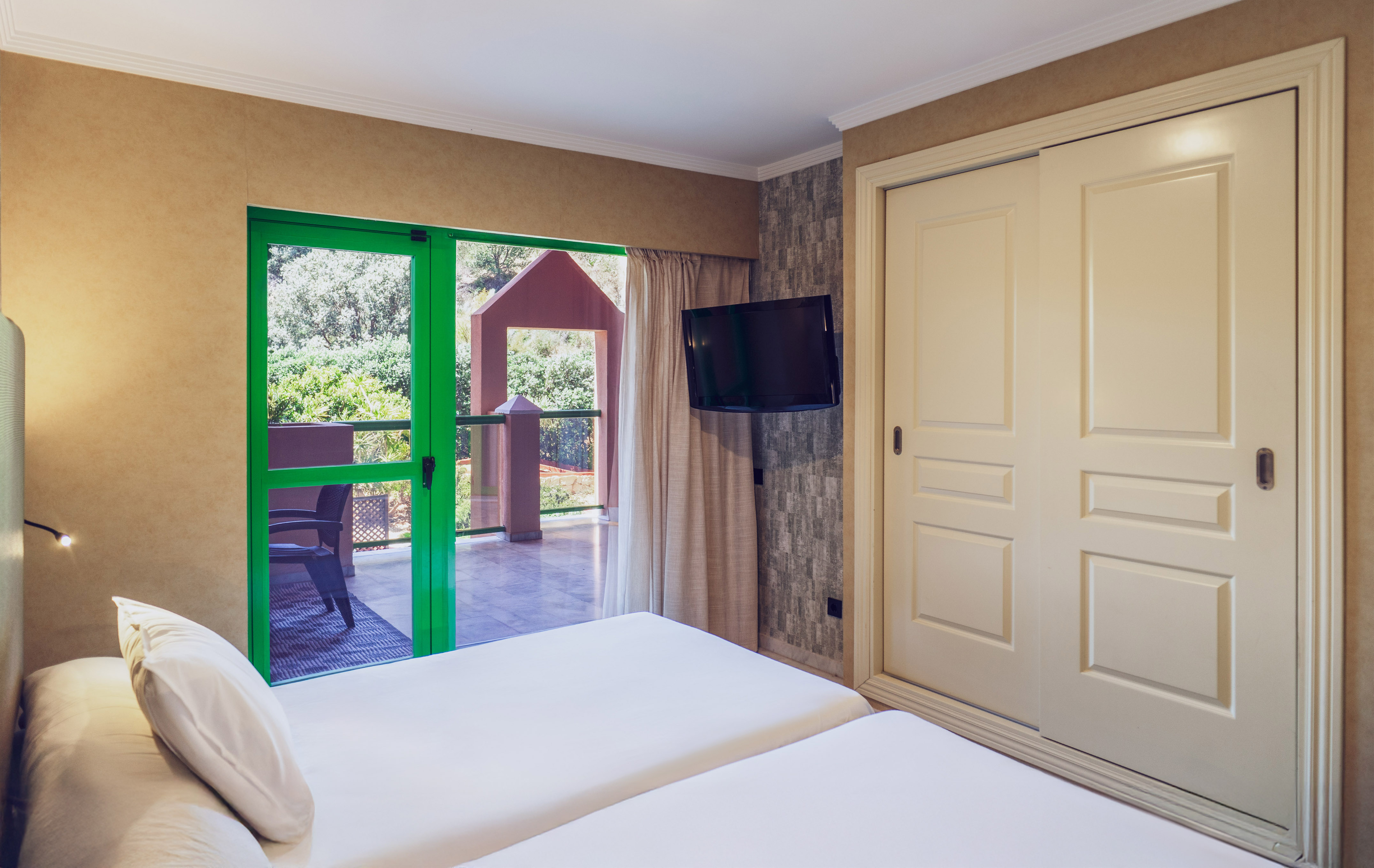 Bright bedroom with twin beds, green balcony doors, wall-mounted TV, and closet.