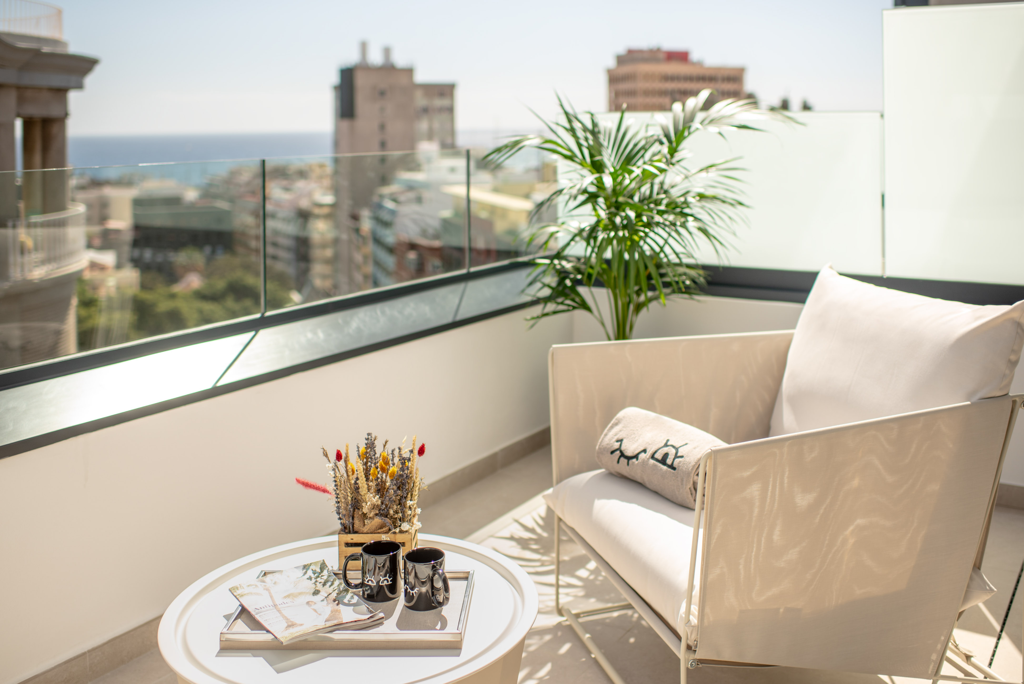 Sunny balcony with urban and sea views, outdoor armchair, coffee table, plant.