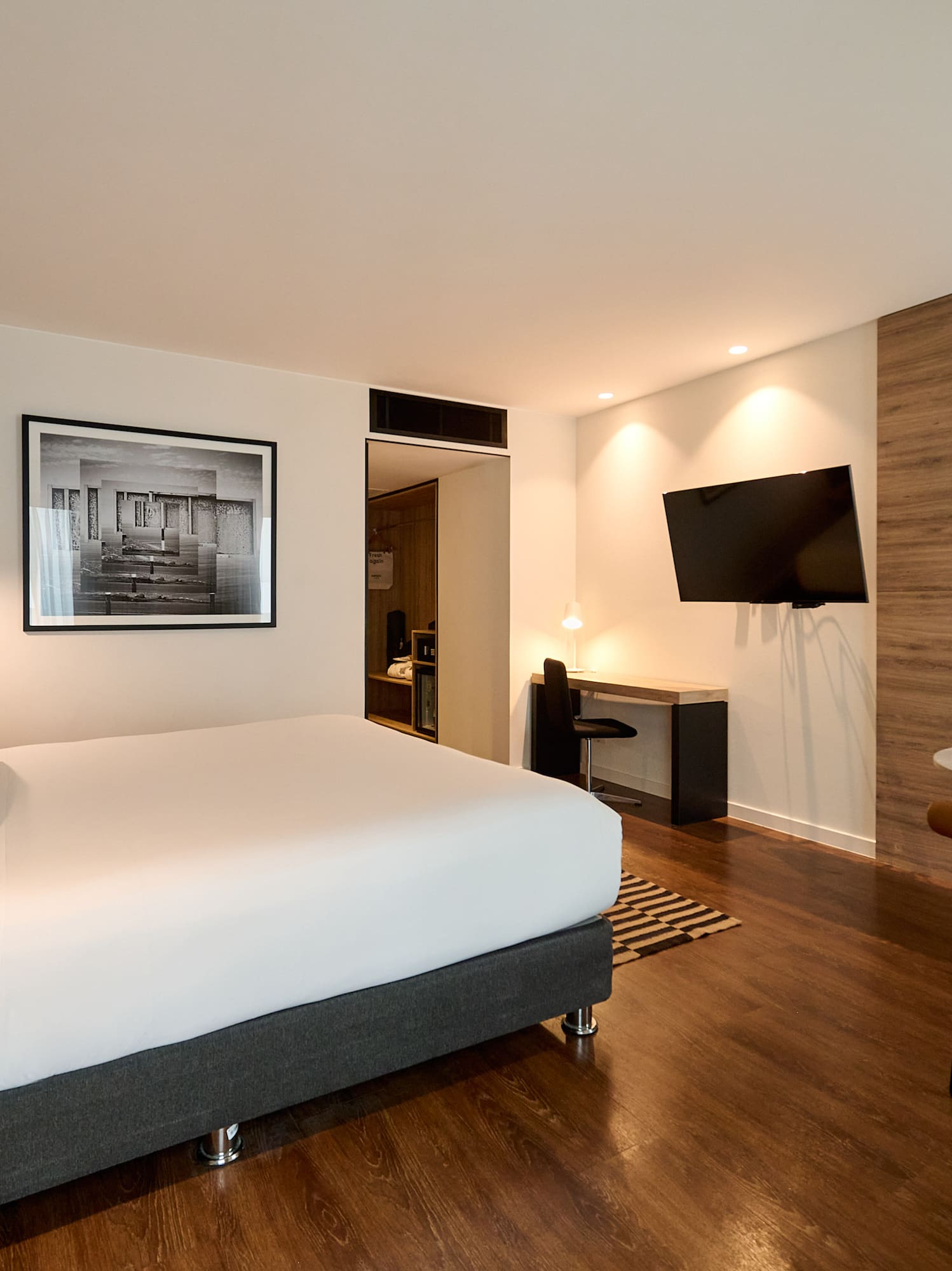 Modern hotel room with king bed, desk, TV, and warm wood flooring.