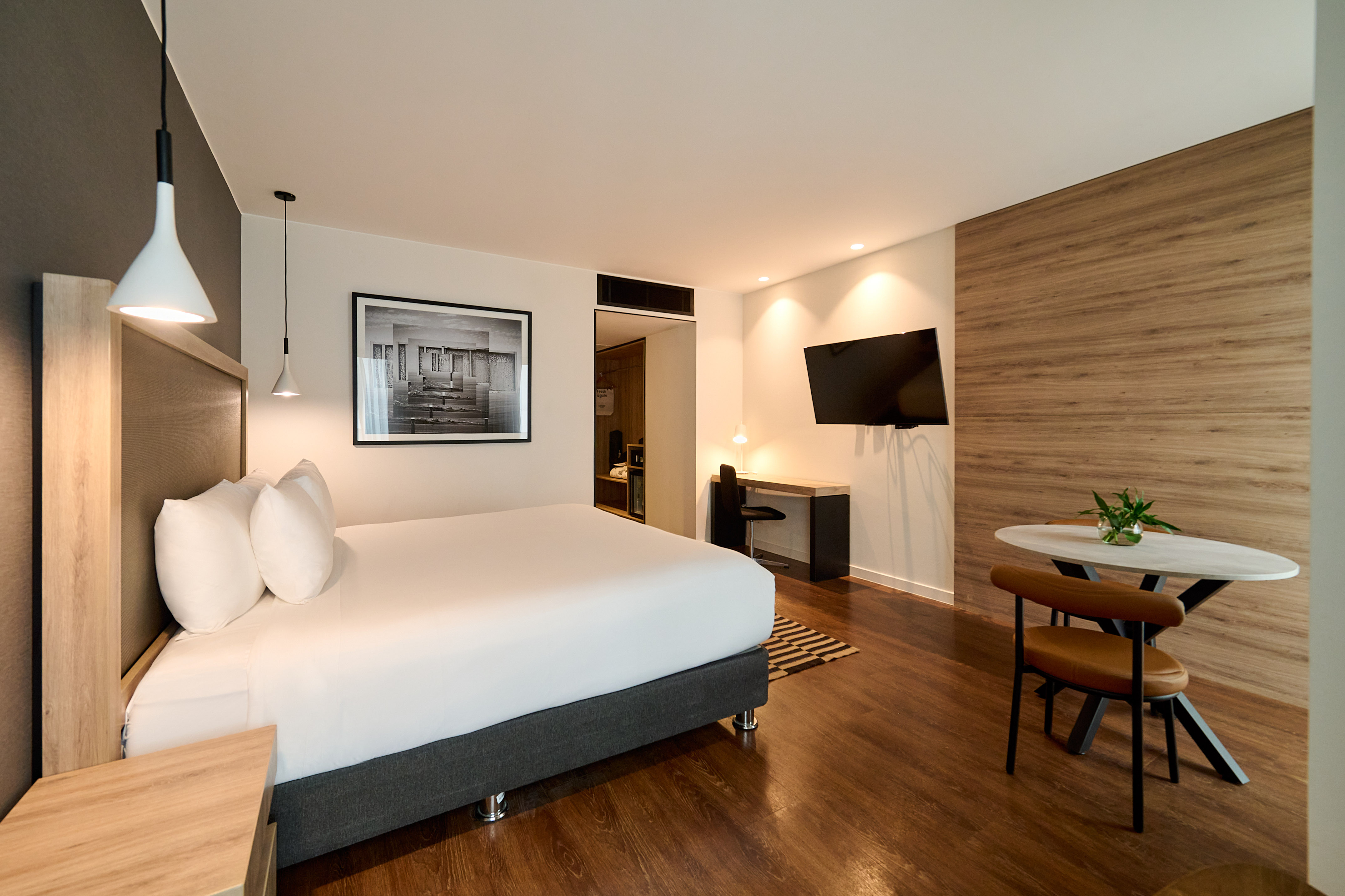 Modern hotel bedroom with white bed, wooden floors, work desk, and dining area.