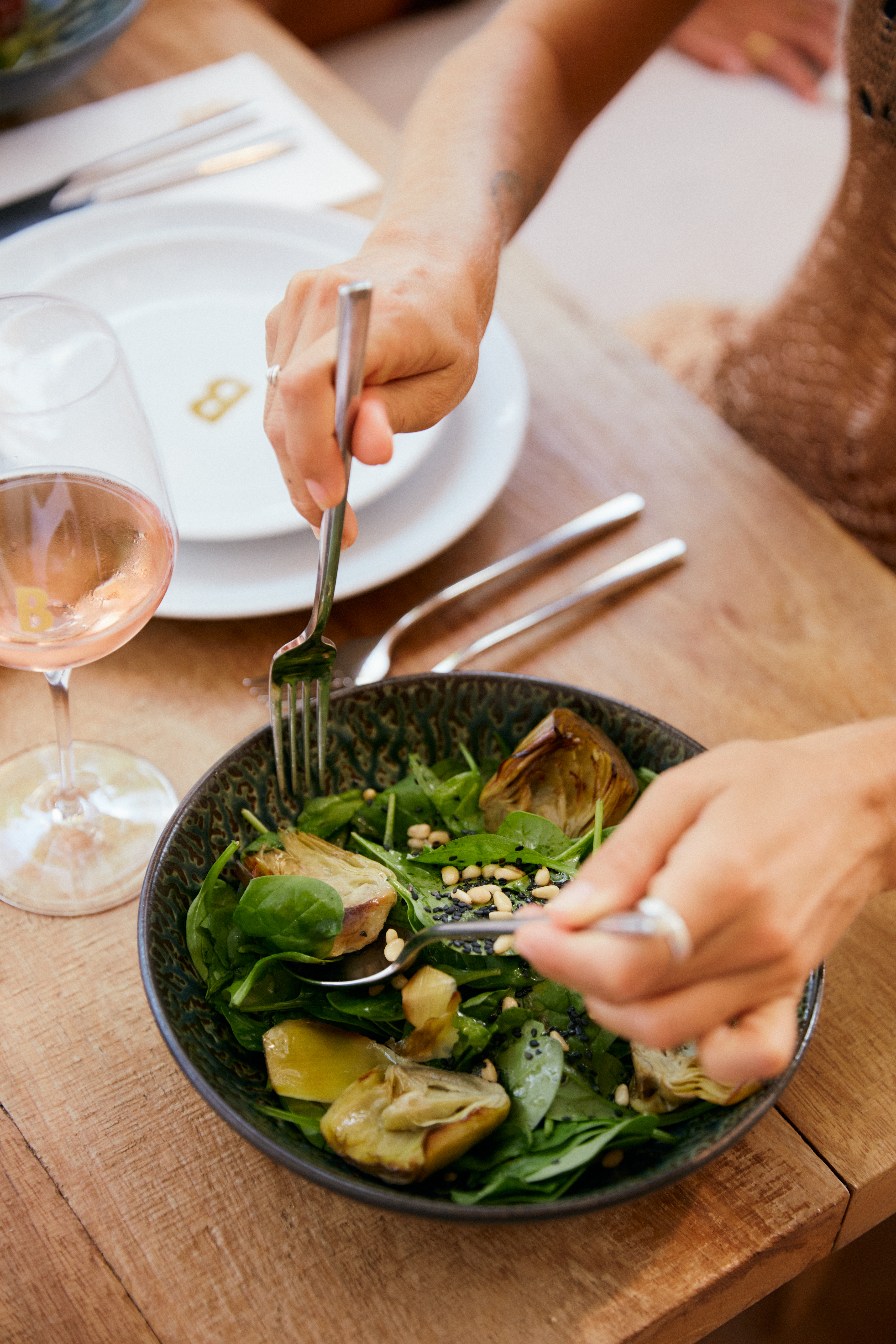 Hands eating spinach artichoke salad with pine nuts, rosé wine nearby.