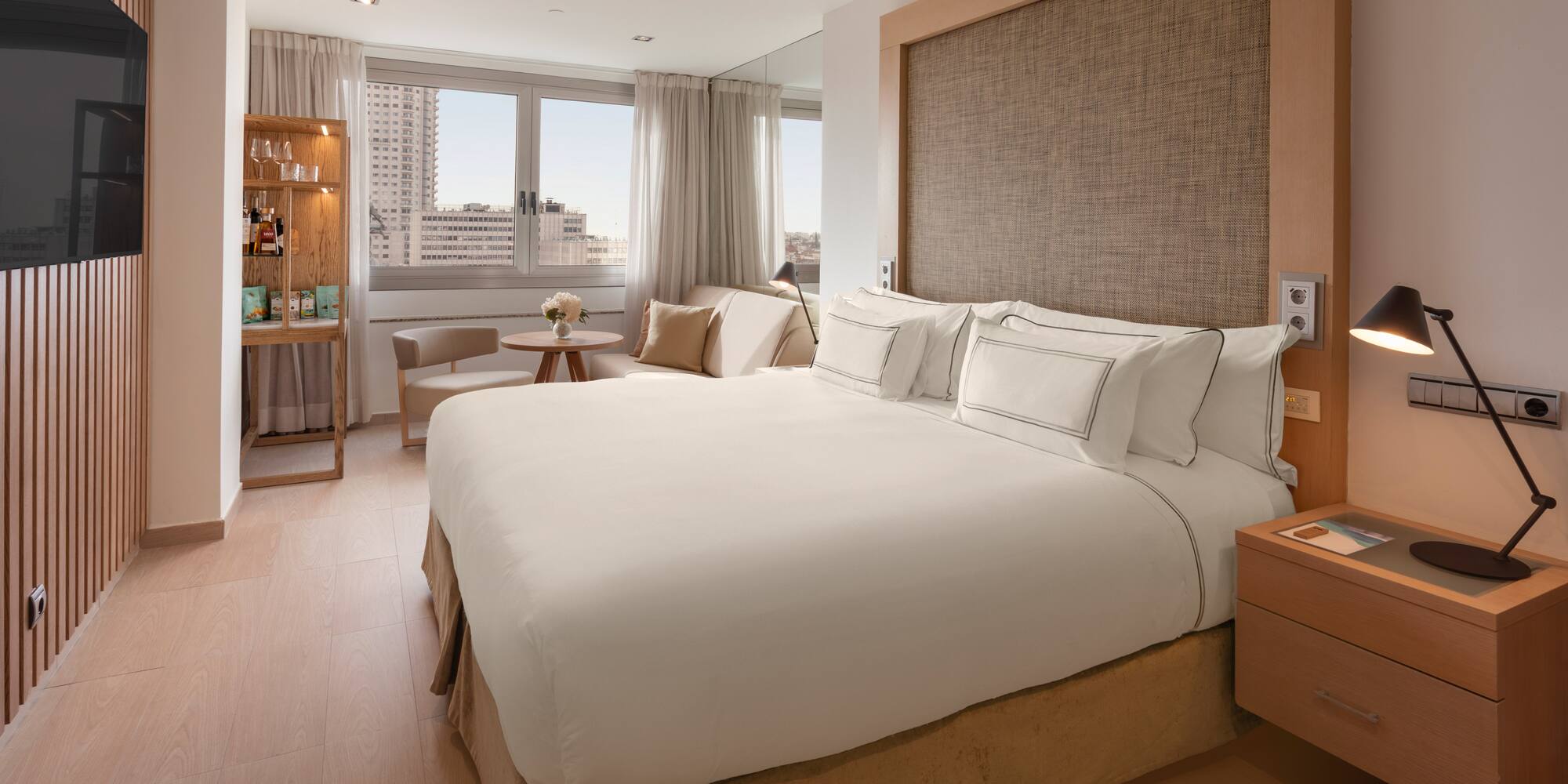 Modern hotel bedroom with white bed, city view, wooden decor, and minibar.