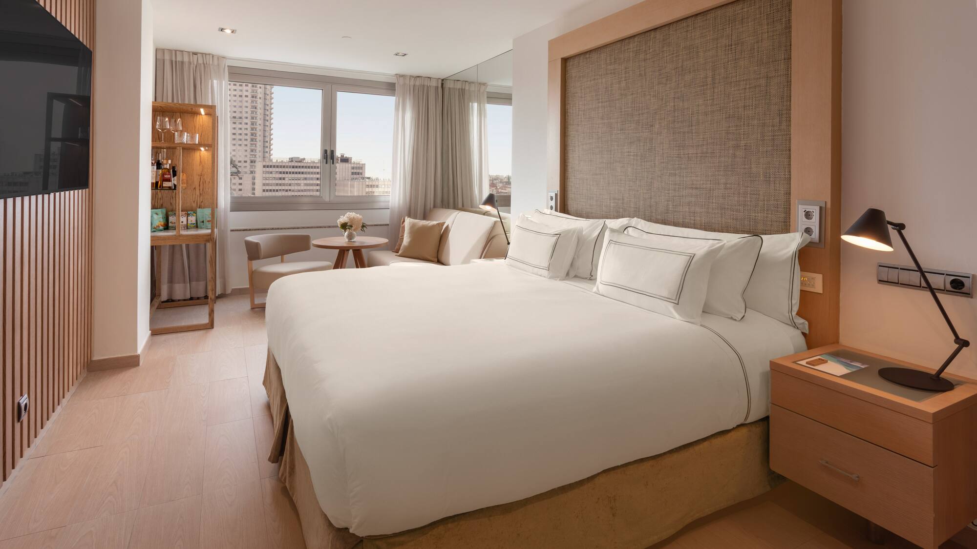 Modern hotel bedroom with white bed, city view, wooden decor, and minibar.