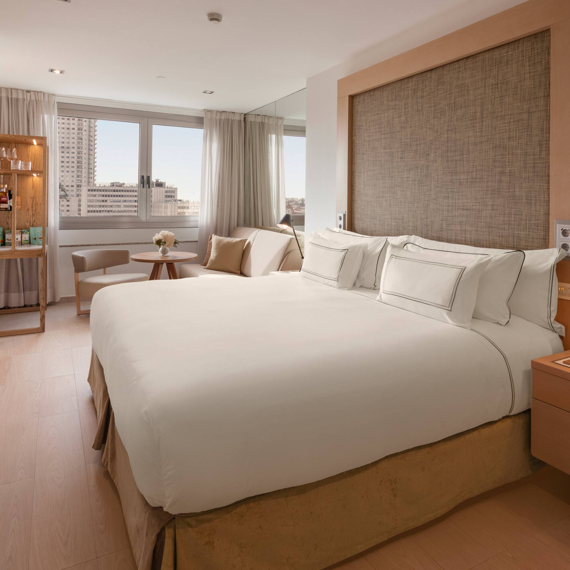Modern hotel bedroom with white bed, city view, wooden decor, and minibar.
