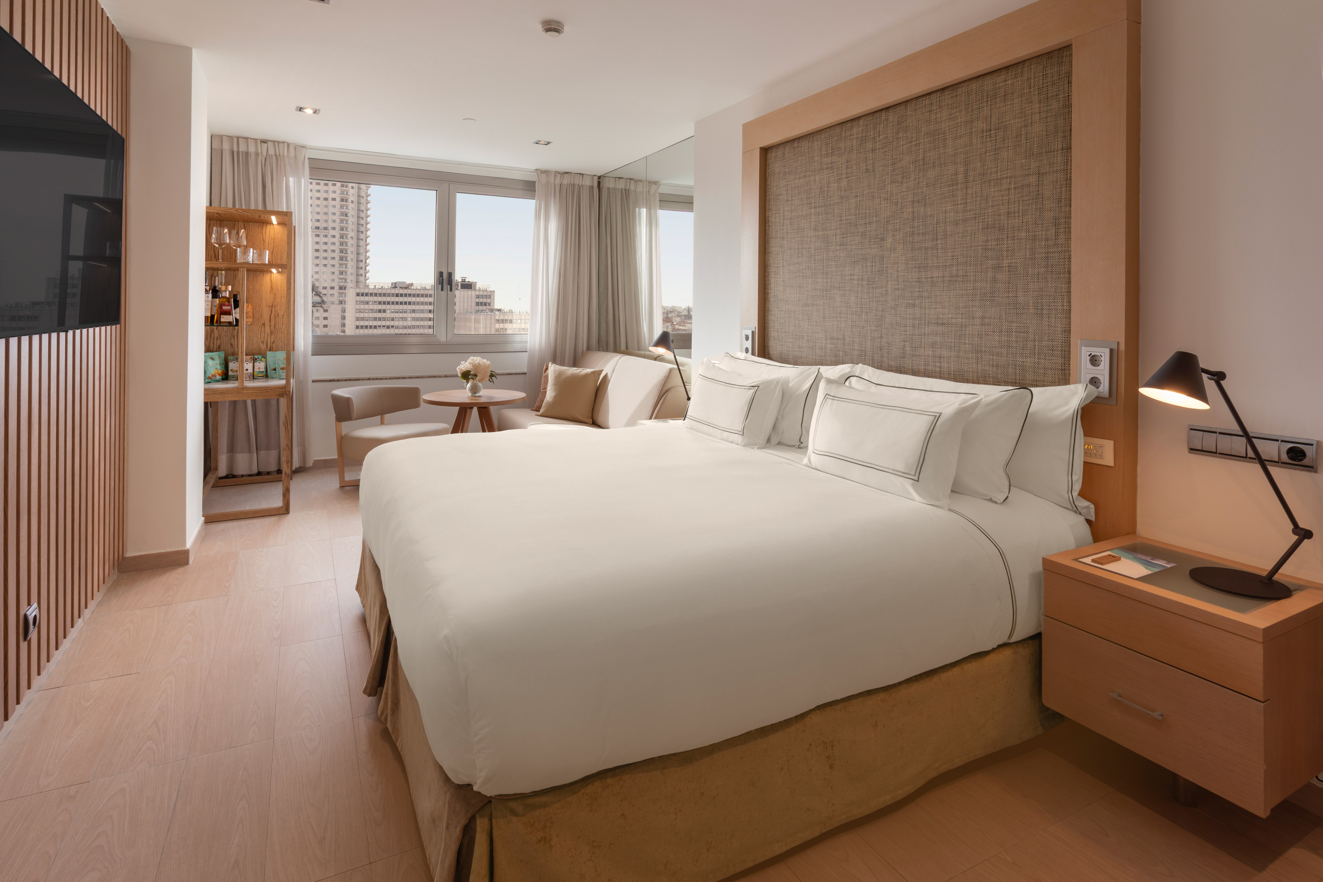 Modern hotel bedroom with king bed, city view, wood accents, and lounge area.