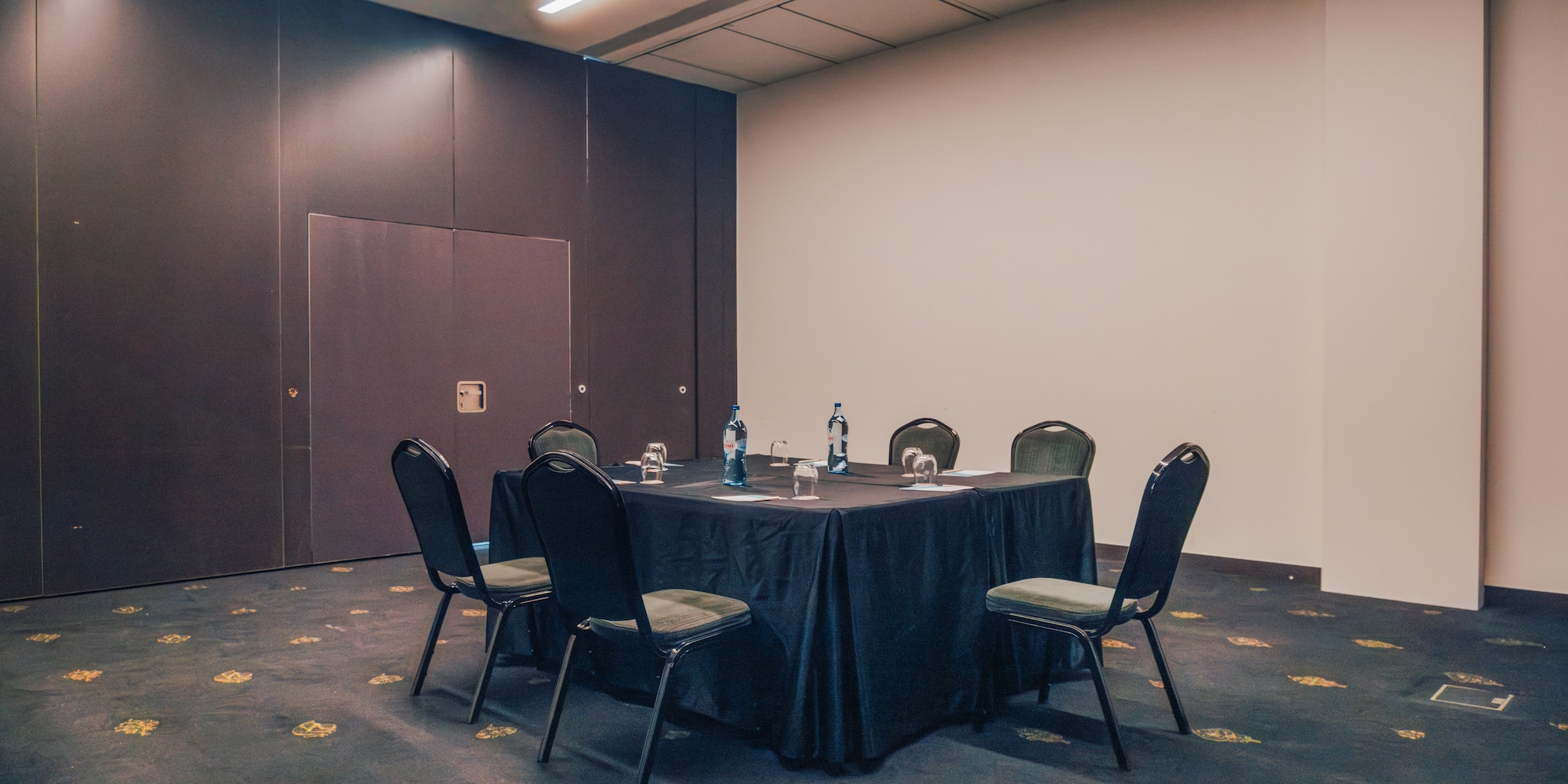Conference room with square table, chairs, water bottles, and dark patterned carpet.