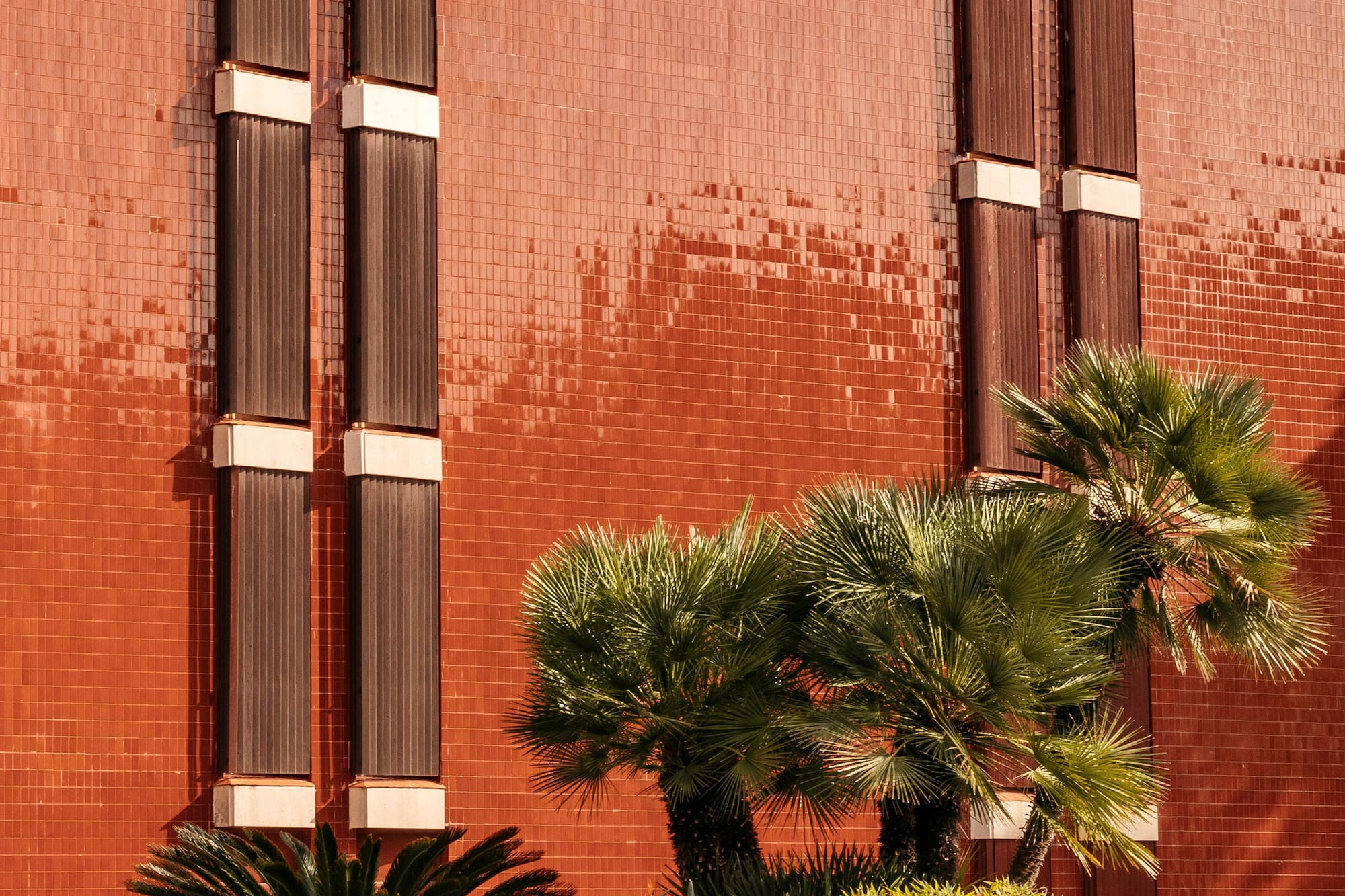 Red tiled facade with brown vertical panels, palm trees, and fountain.