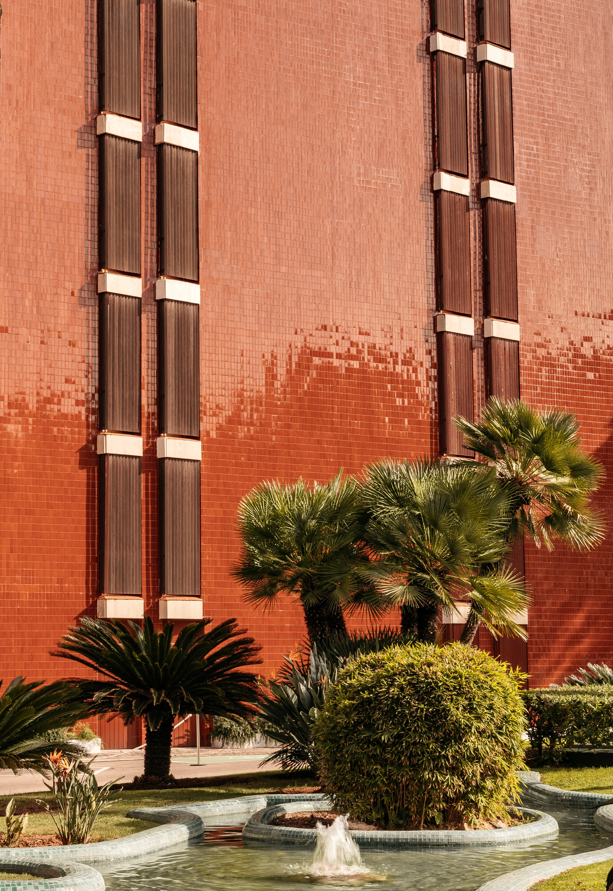 Red tiled facade with brown vertical panels, palm trees, and fountain.