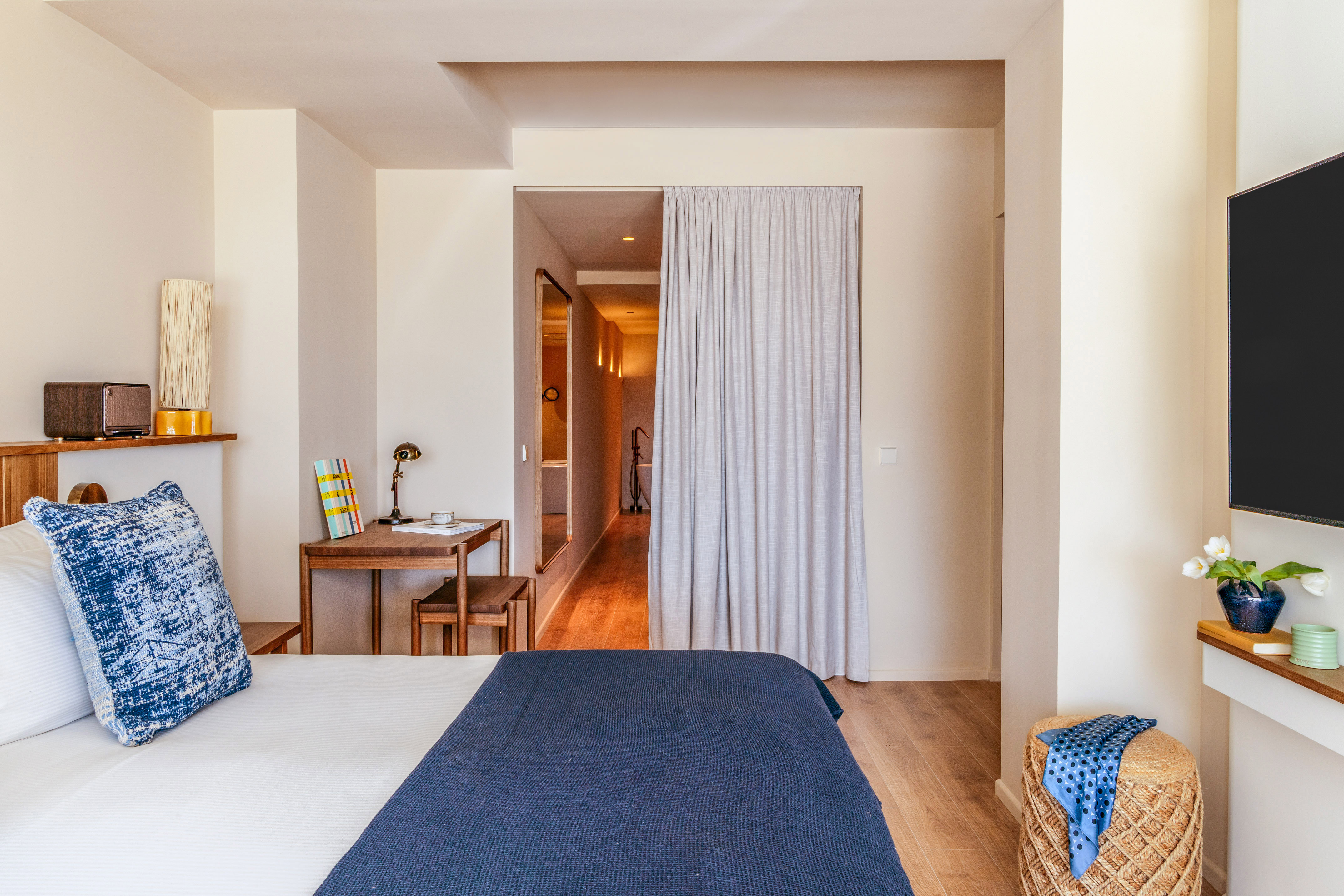Modern hotel room with blue and white bed, wooden desk, and adjoining hall.