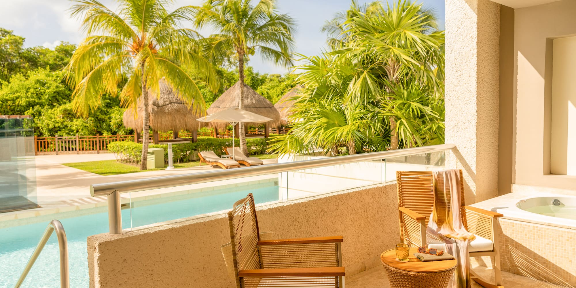 Sunny tropical balcony with private jacuzzi, pool, palm trees, and resort cabanas.