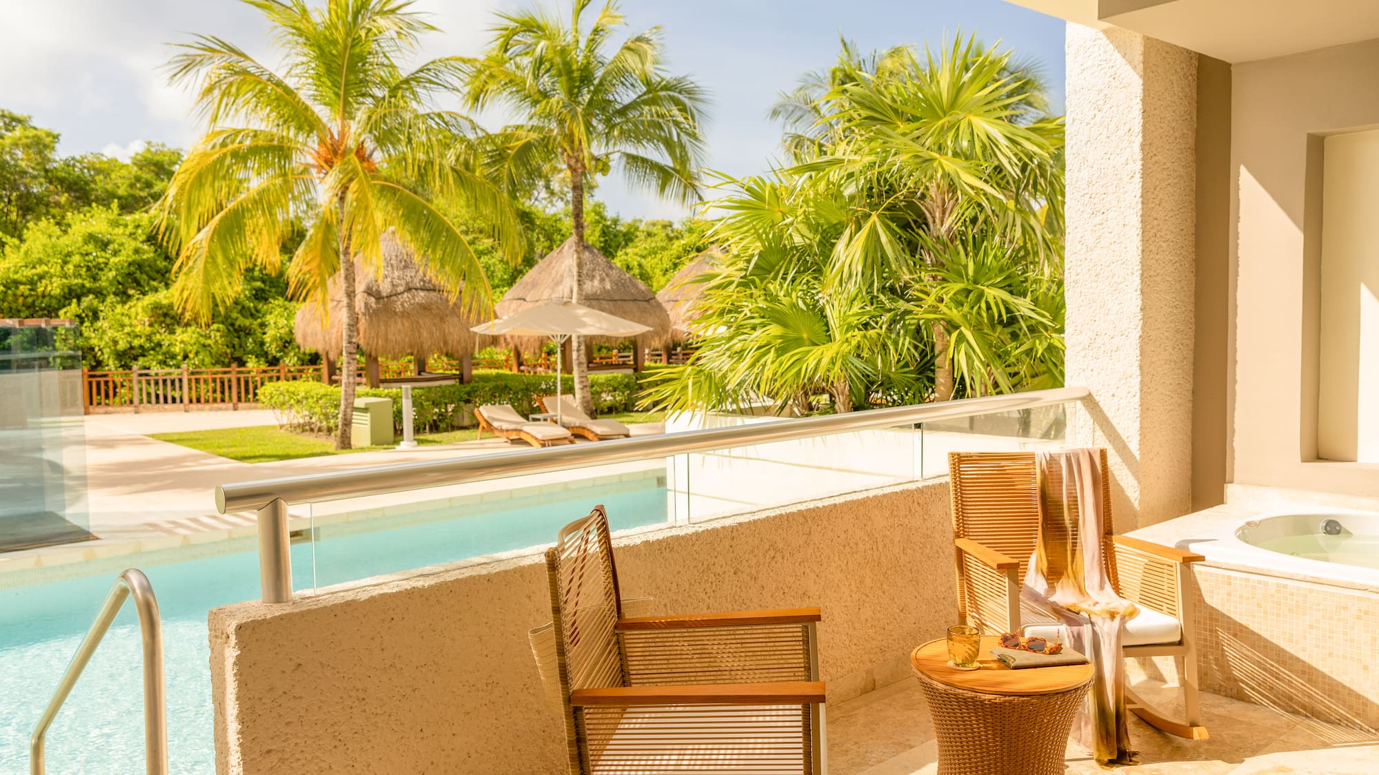 Sunny tropical balcony with private jacuzzi, pool, palm trees, and resort cabanas.