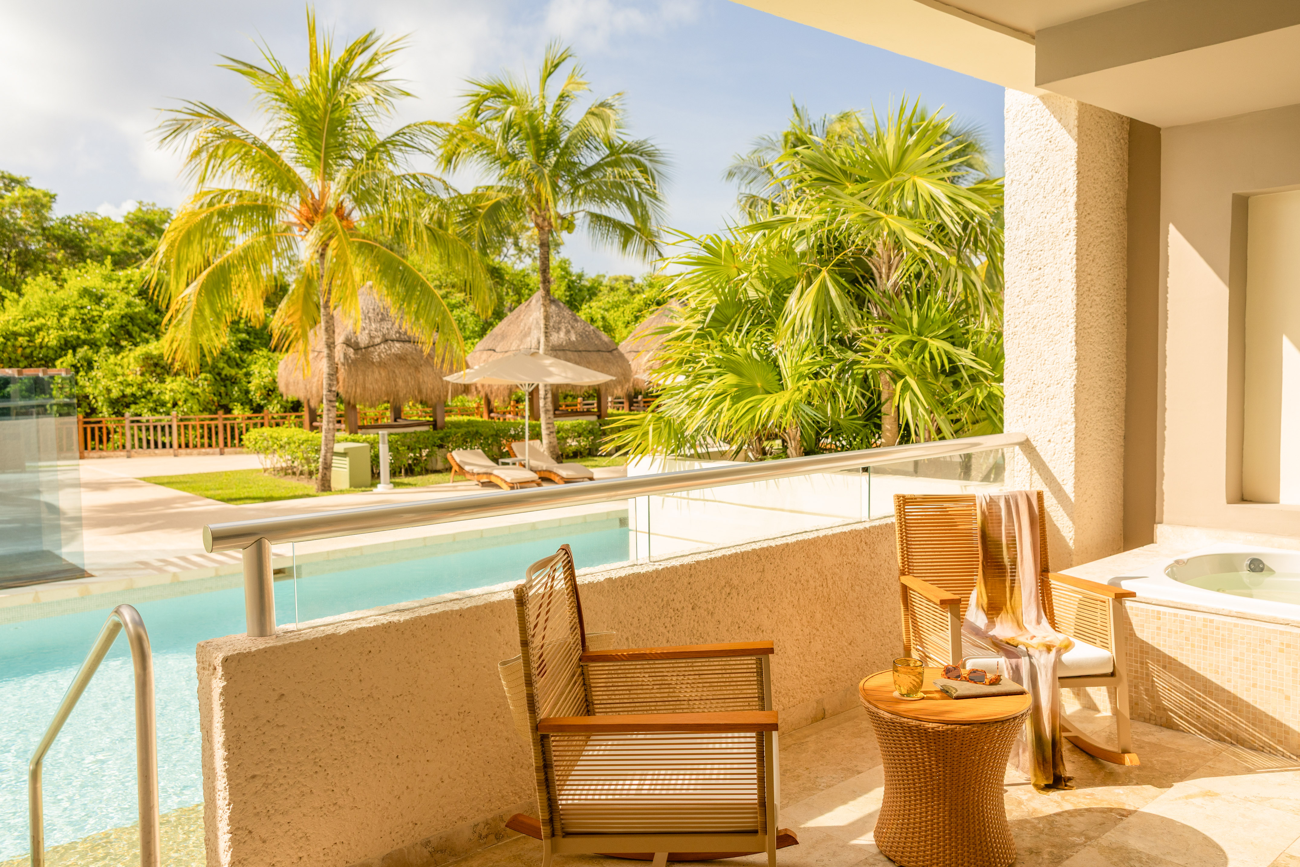 Sunny tropical balcony with private jacuzzi, pool, palm trees, and resort cabanas.