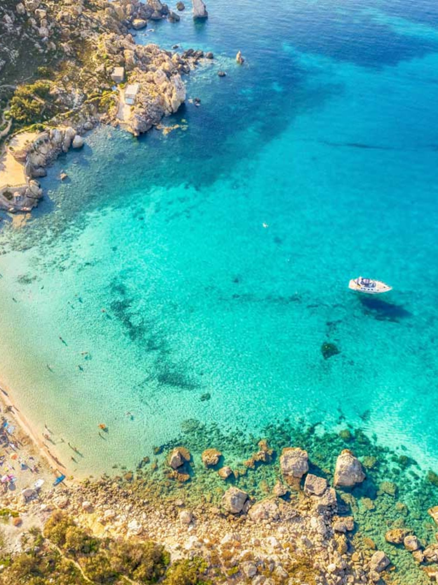 Aerial view of a sandy beach with clear turquoise sea, rocky coast, boats, and resort.