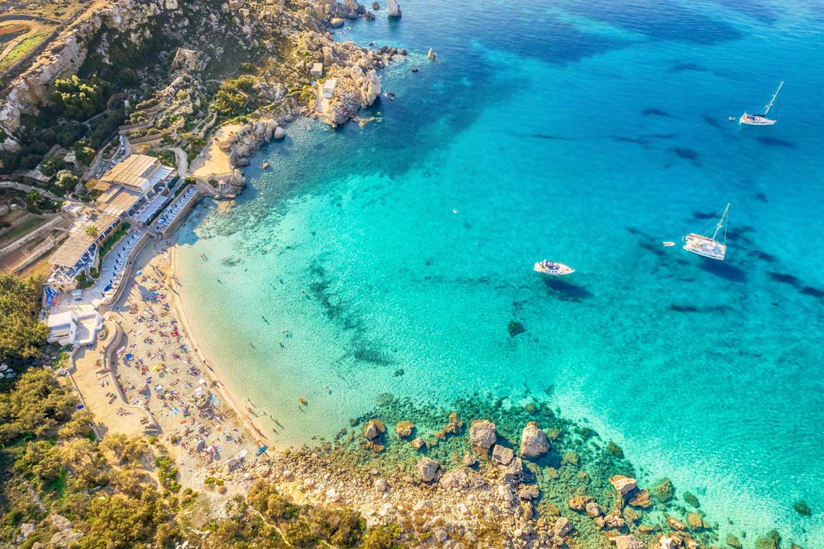 Aerial view of a sandy beach with clear turquoise sea, rocky coast, boats, and resort.