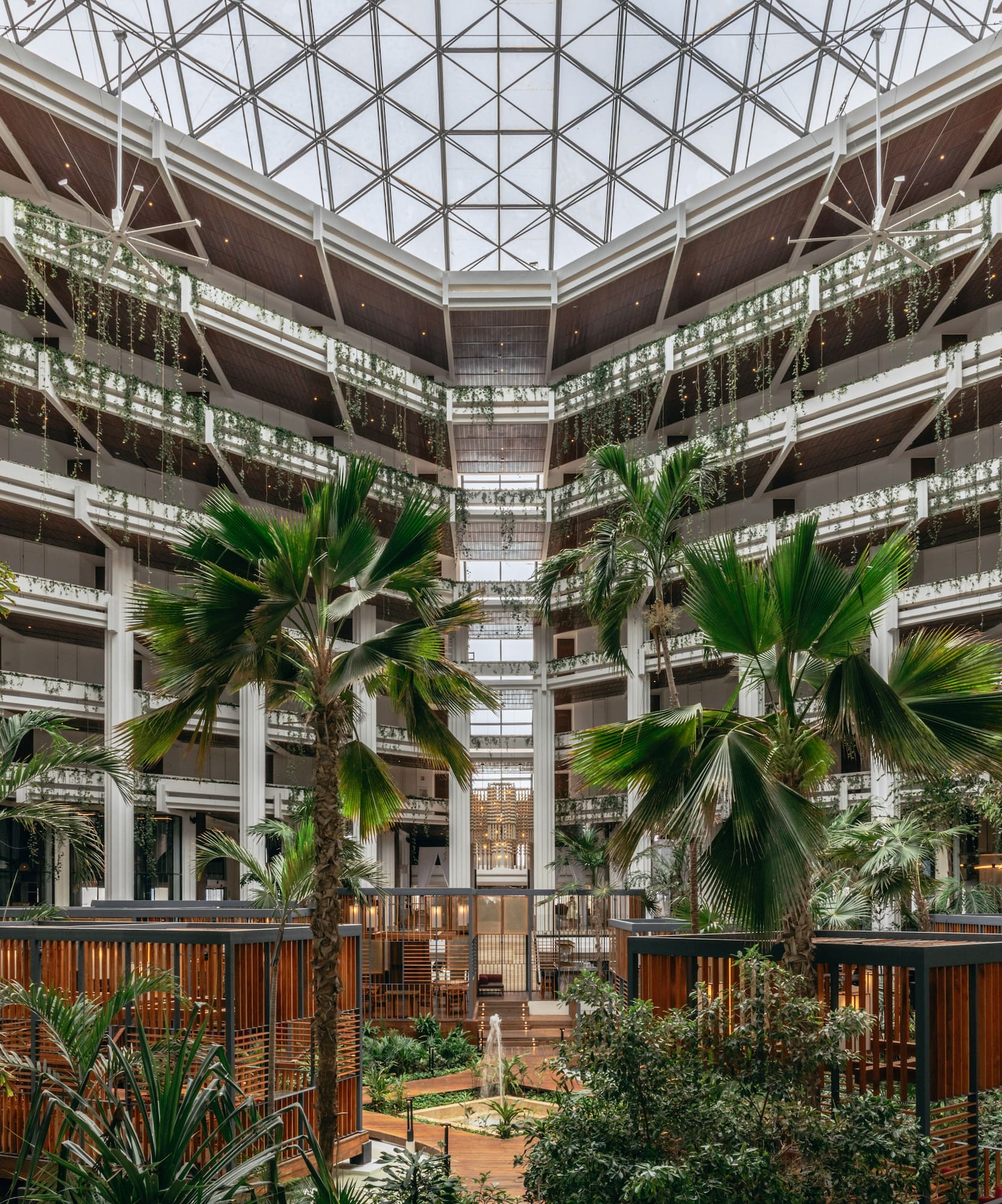 Spacious hotel atrium with tropical plants, wooden details, and glass ceiling.