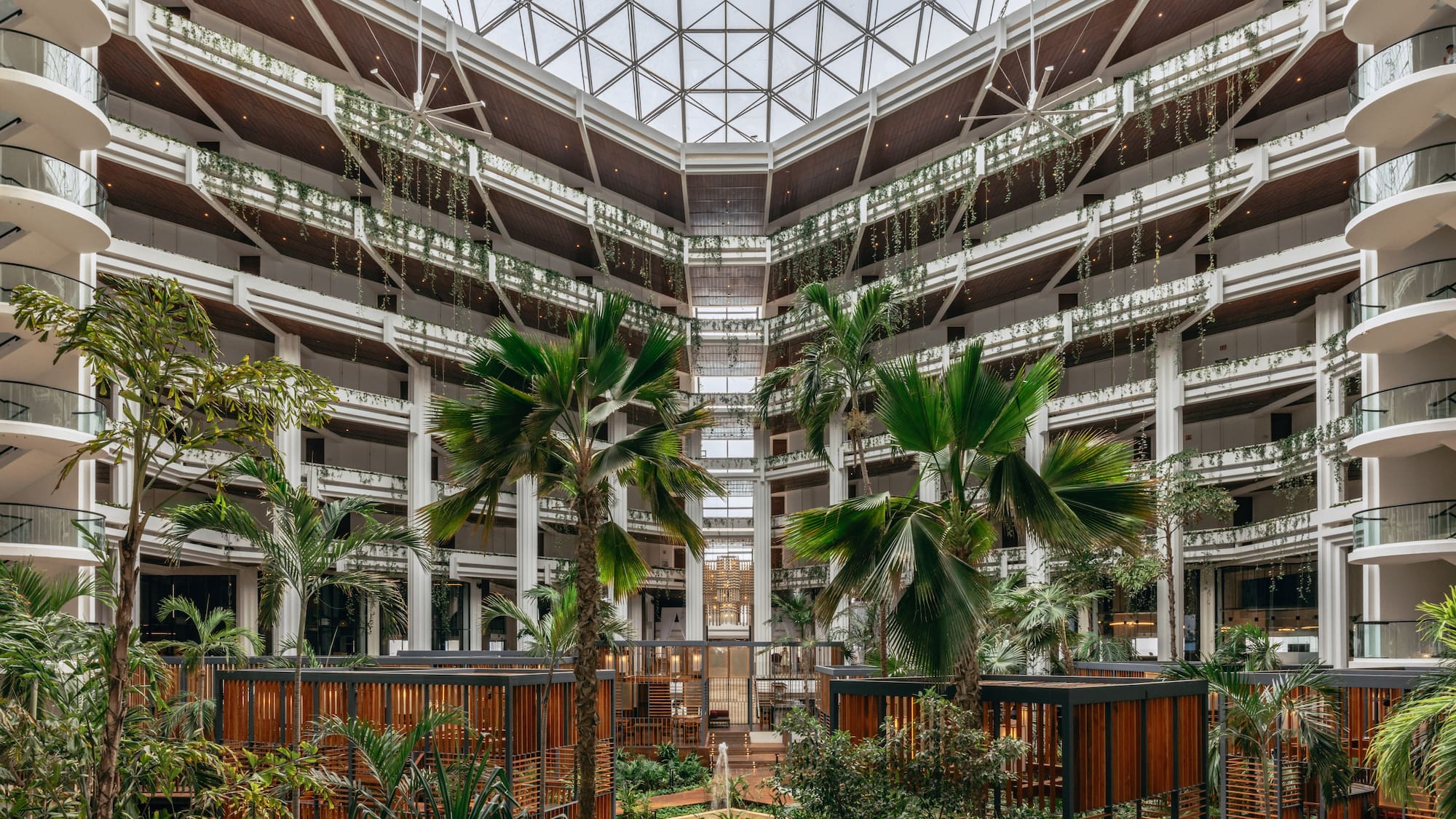 Spacious hotel atrium with glass roof, tropical plants, and modern balconies.