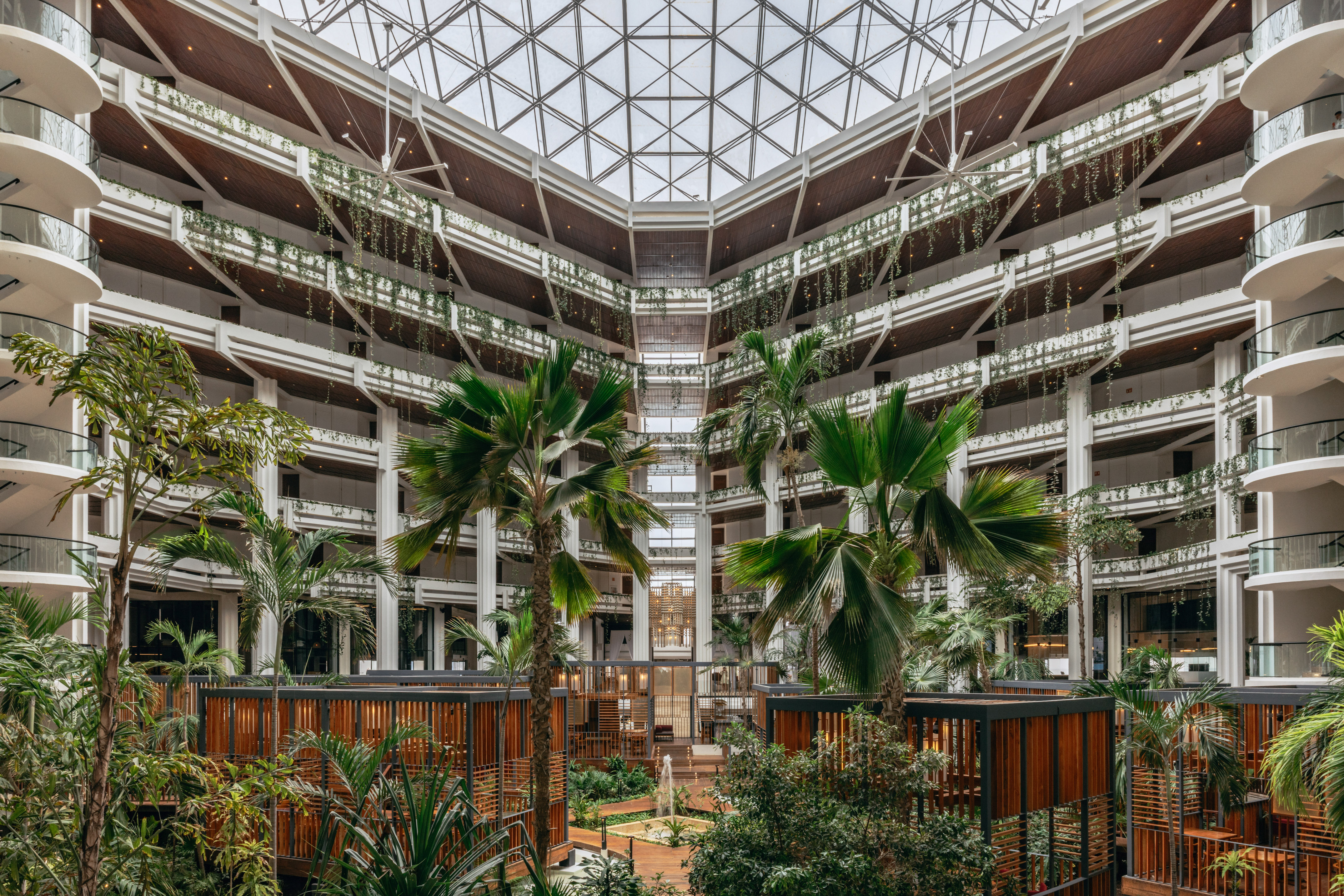 Spacious hotel atrium with tropical plants, wooden details, and glass ceiling.