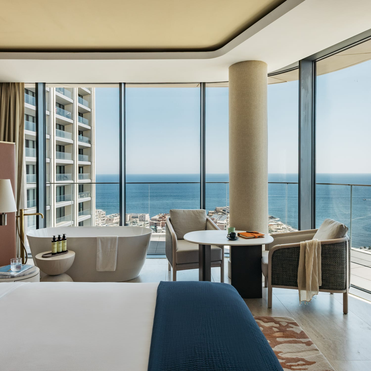 Modern hotel suite with ocean view, bed, freestanding tub, and large windows.