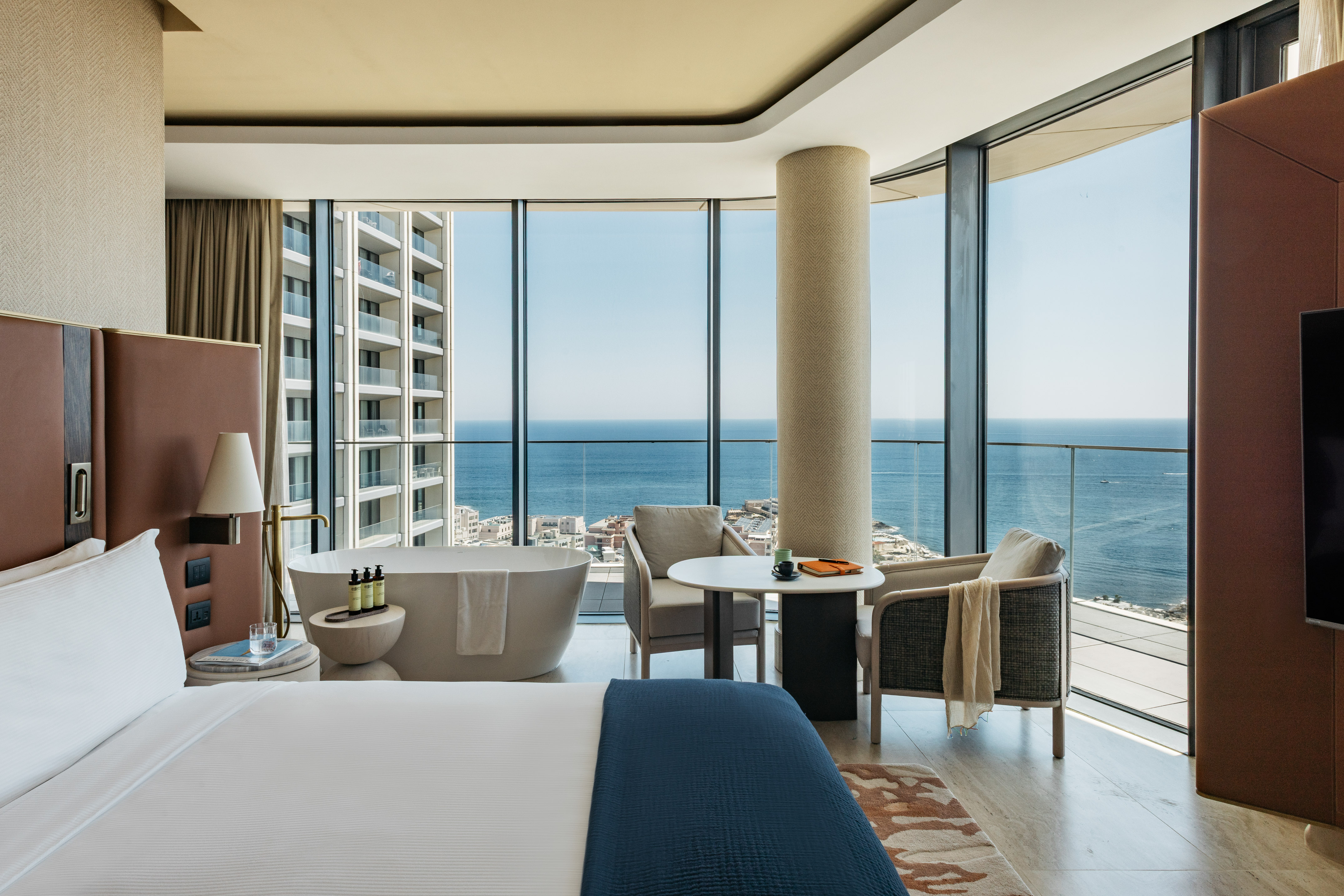 Modern hotel suite with ocean view, bed, freestanding tub, and large windows.