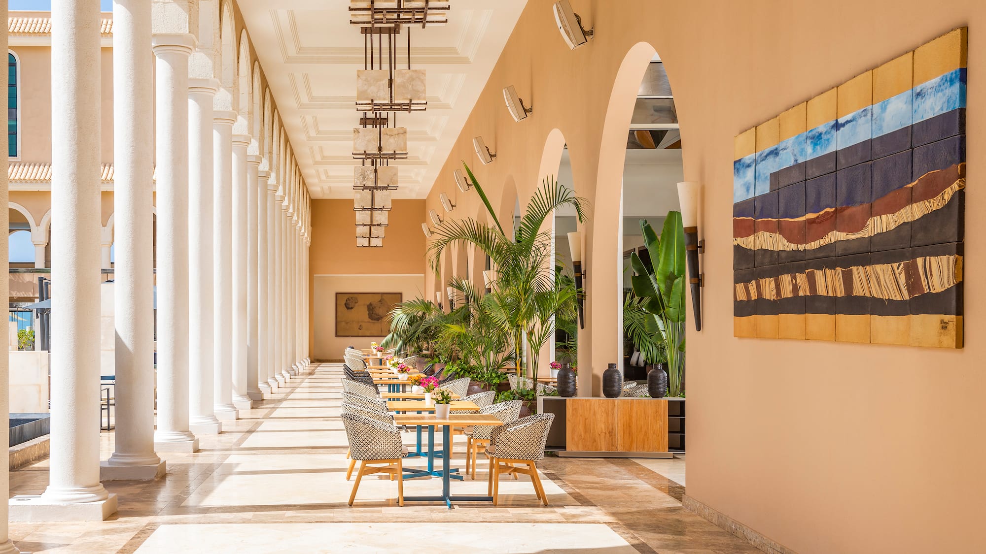 Resort patio with white columns, outdoor tables, tropical plants, and abstract art.