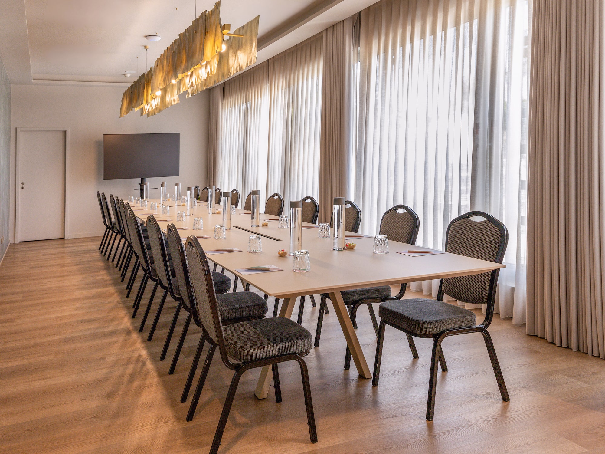 Modern conference room with long table, many chairs, large TV, and bright curtains.