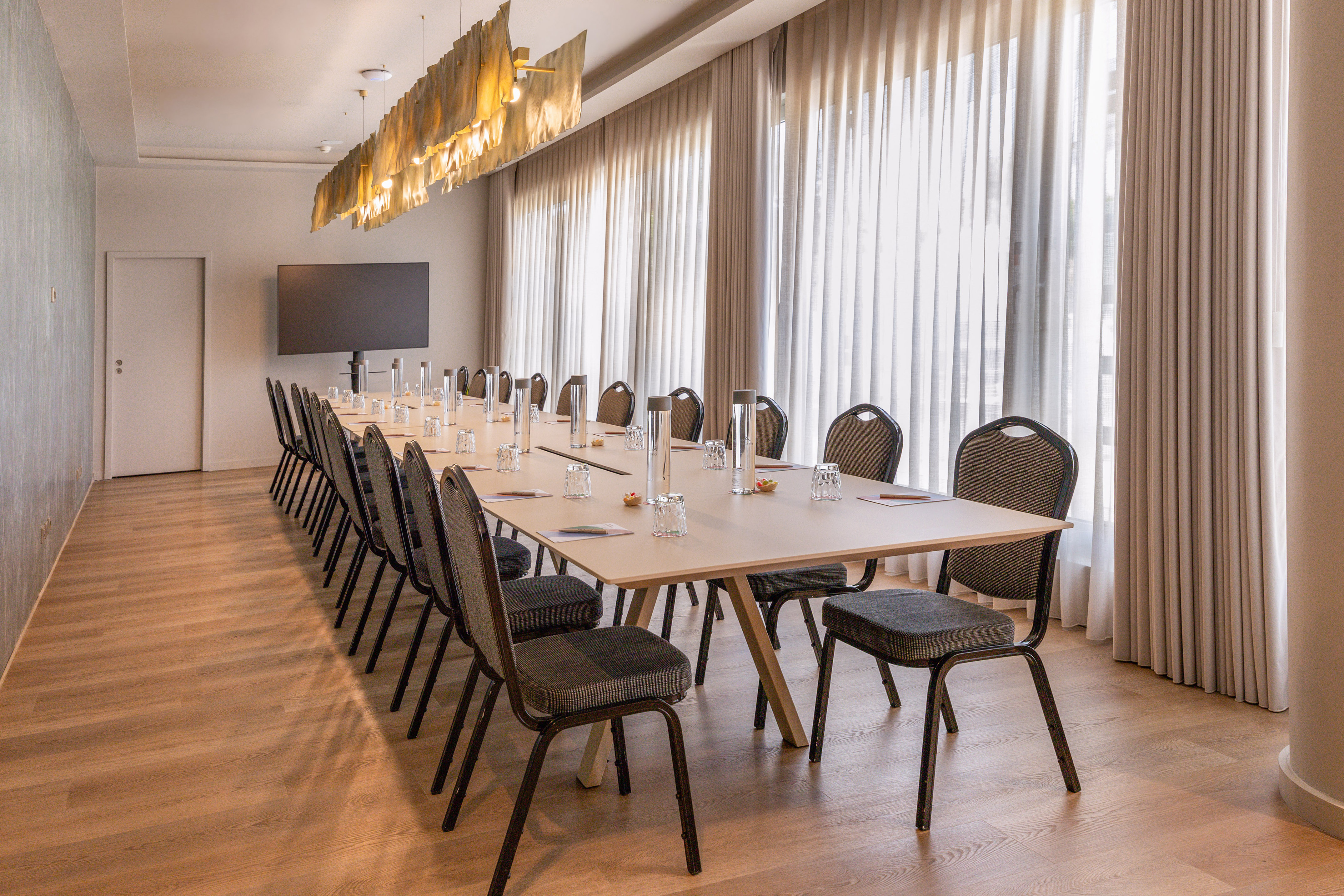 Modern conference room with long table, many chairs, large TV, and bright curtains.