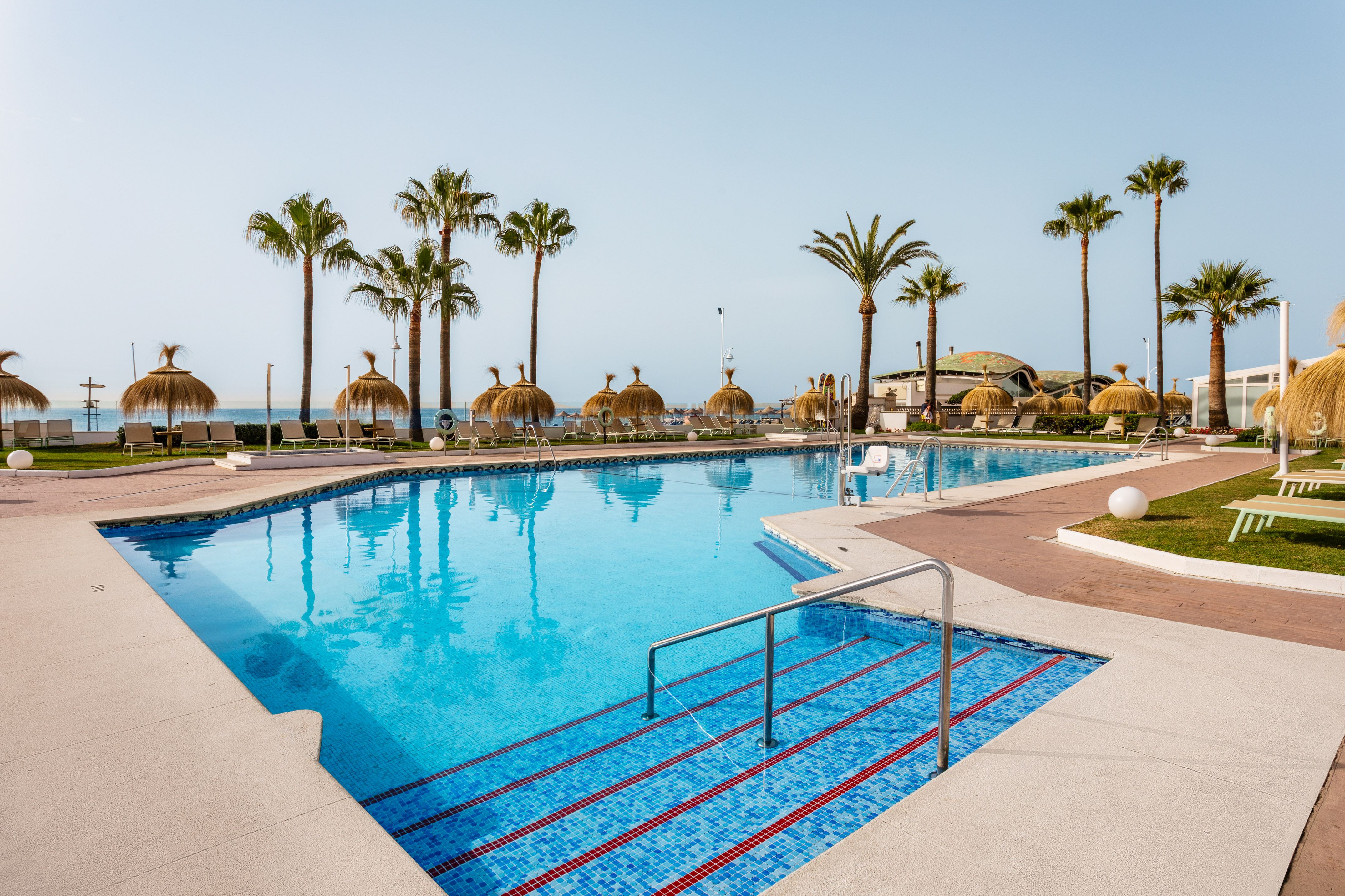 Tropical resort pool with palm trees, straw parasols, and distant ocean.