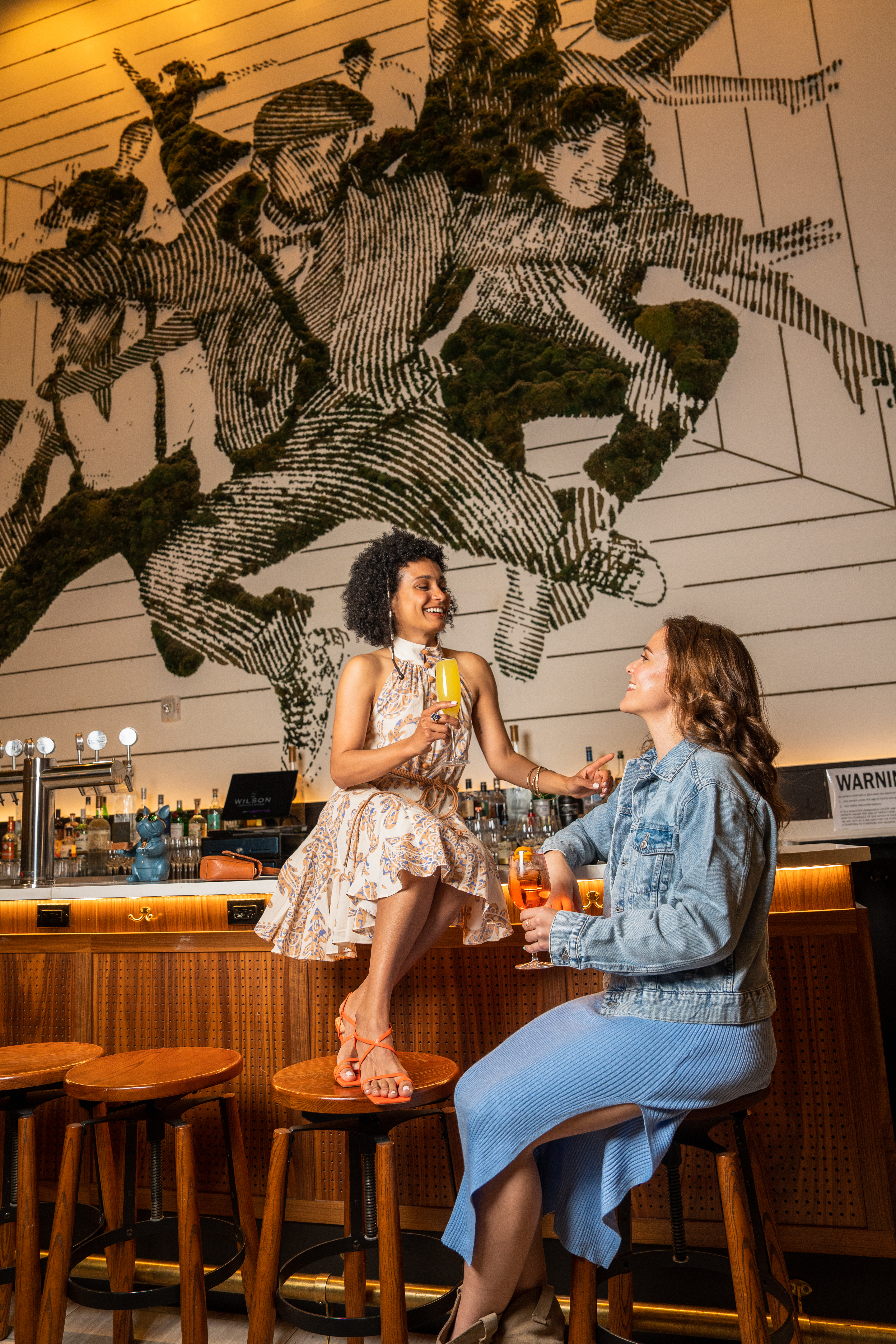 Two women laughing, enjoying cocktails at a vibrant bar with a green moss mural.