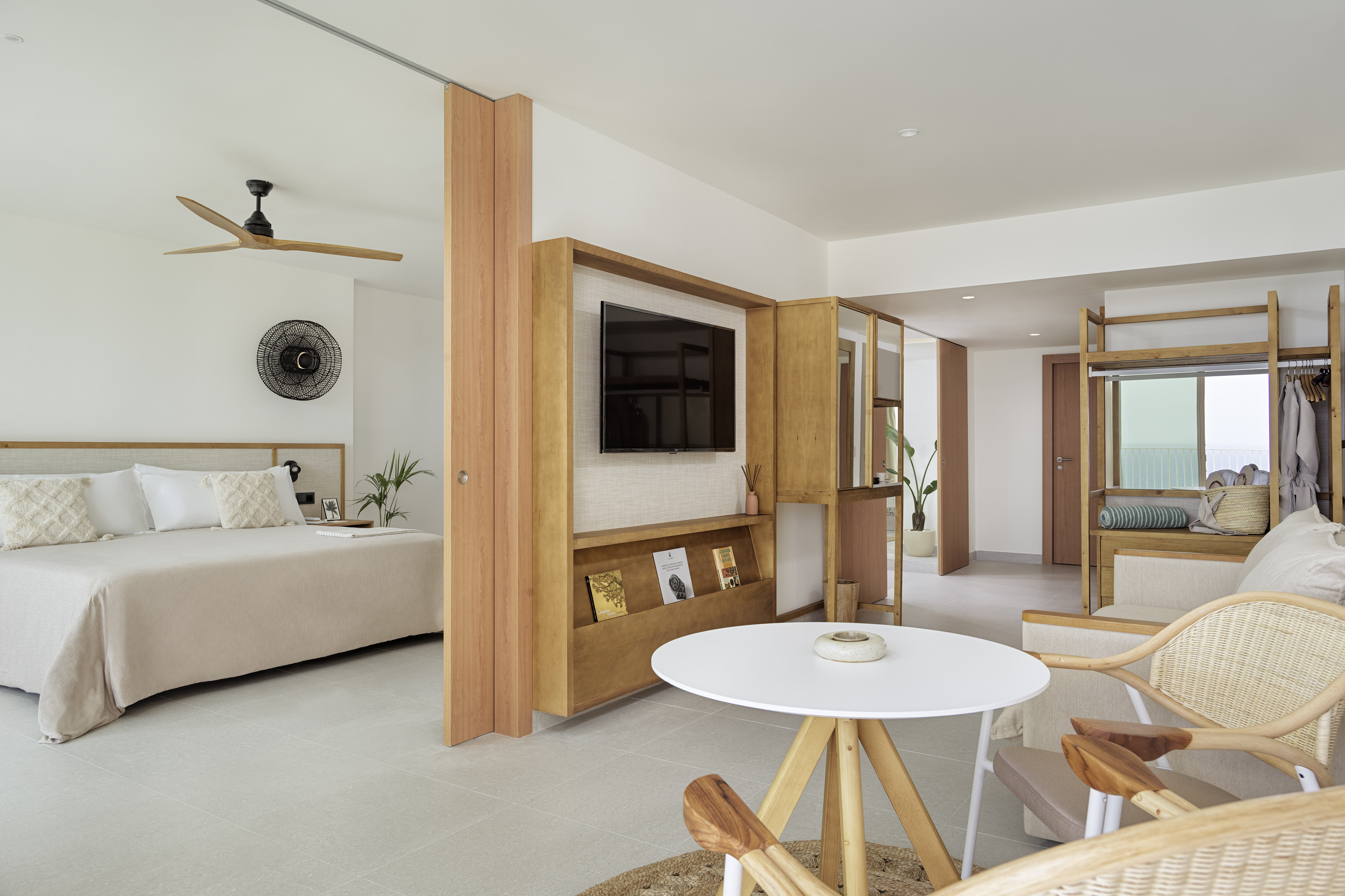 Modern hotel suite with bedroom, sitting area, wooden TV unit, and light decor.