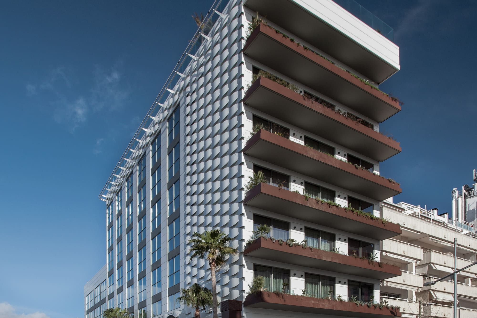 Modern hotel with patterned facade, balconies, palm trees, and blue sky.