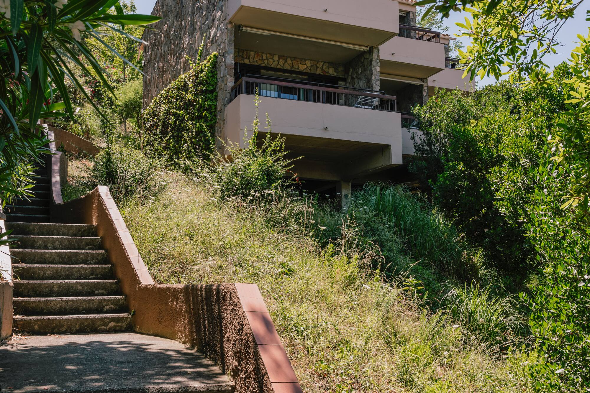 Hillside stone building with balconies above concrete stairs and lush greenery.