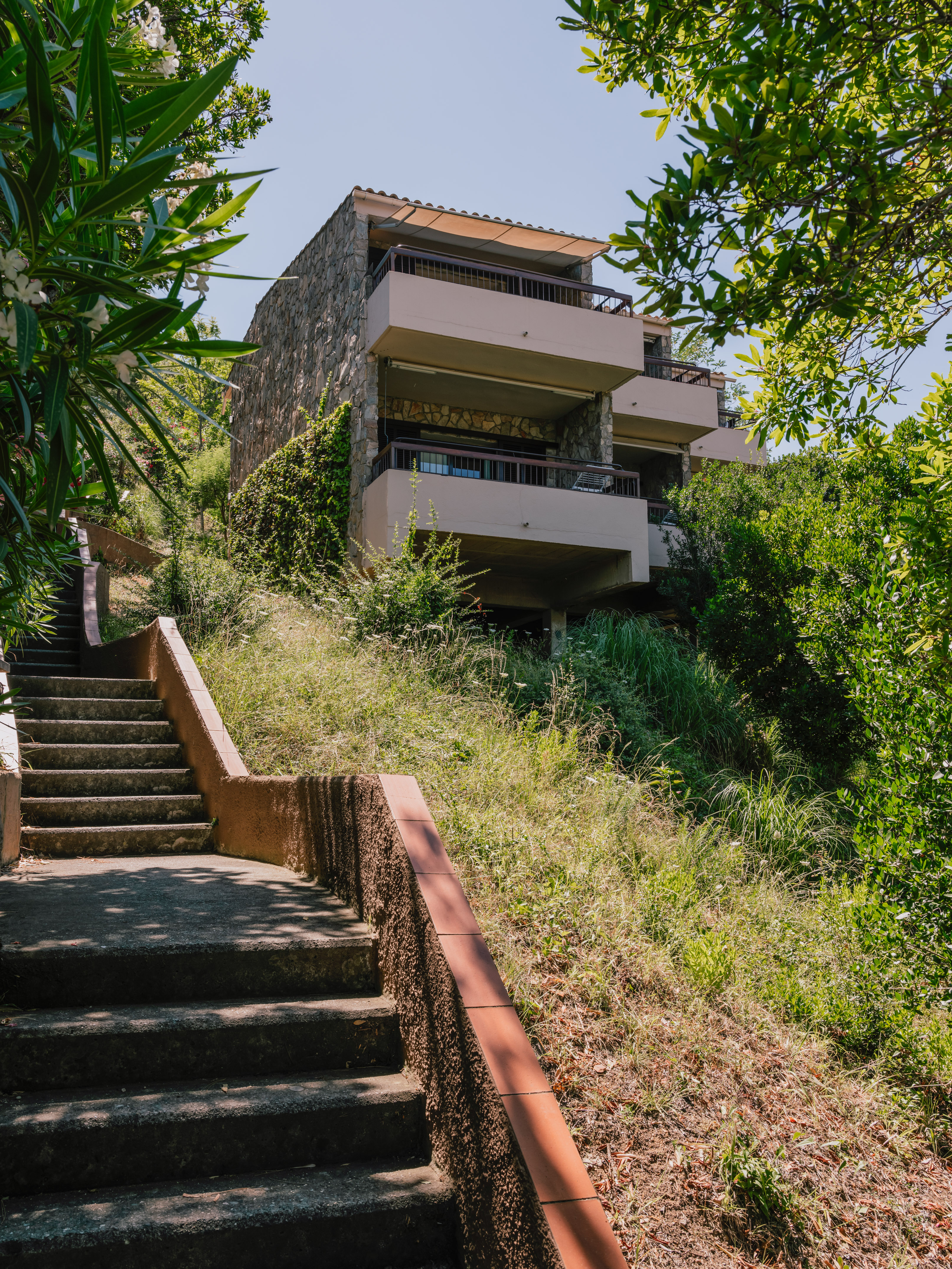 Hillside stone building with balconies above concrete stairs and lush greenery.