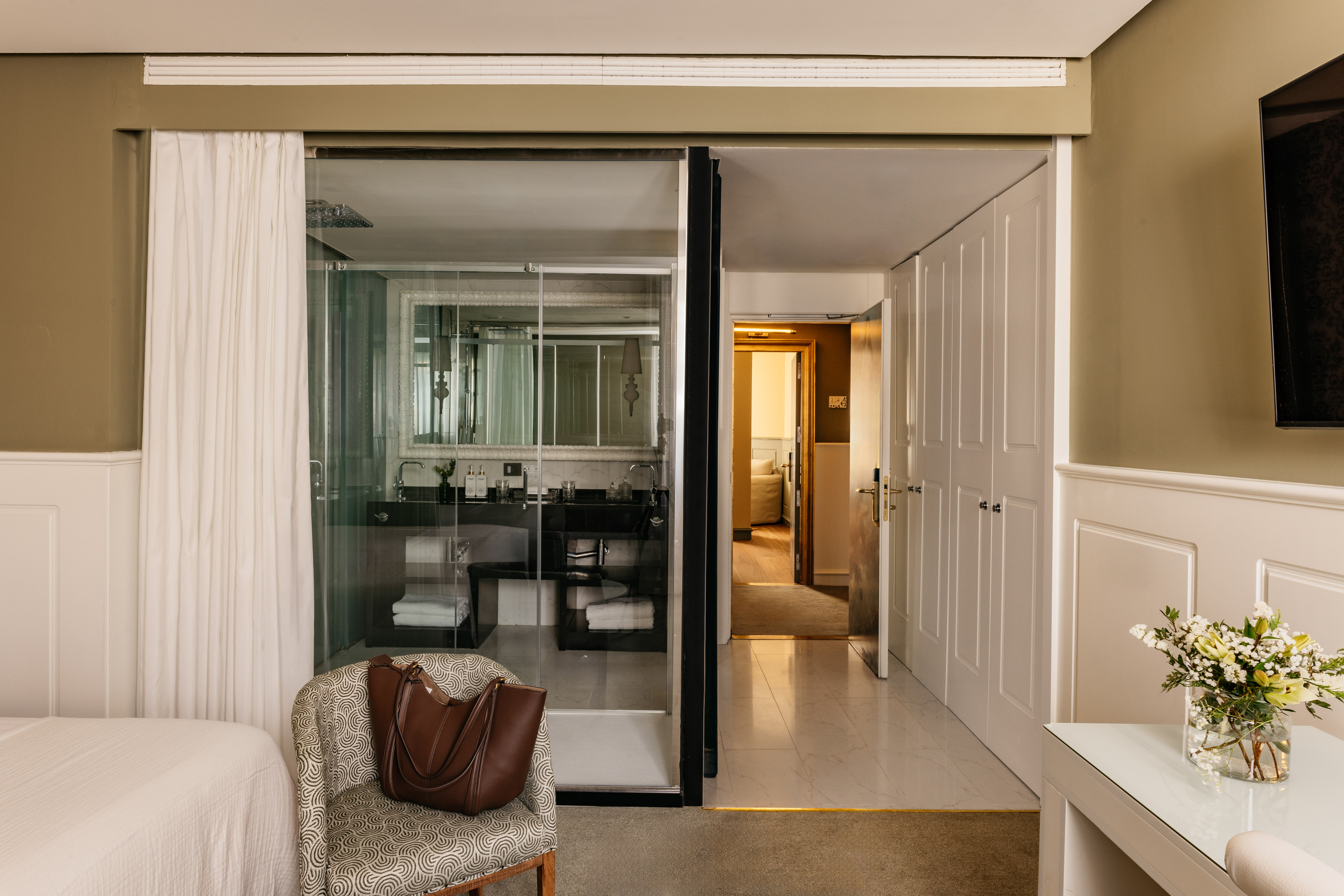 Suite de hotel elegante con baño de cristal, dormitorio y recibidor.