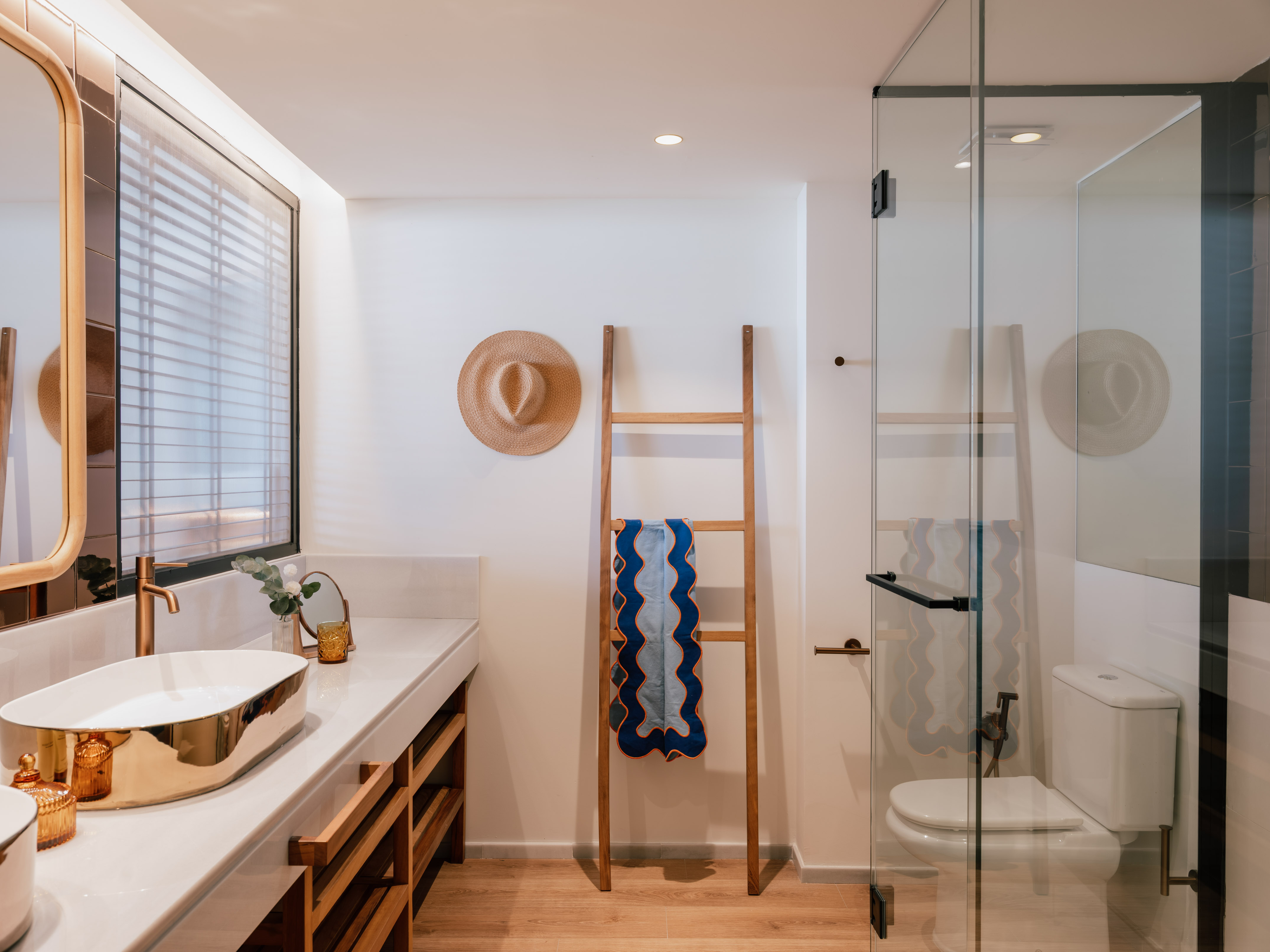 Modern bathroom with gold vessel sink, wood vanity, glass shower, and straw hat.