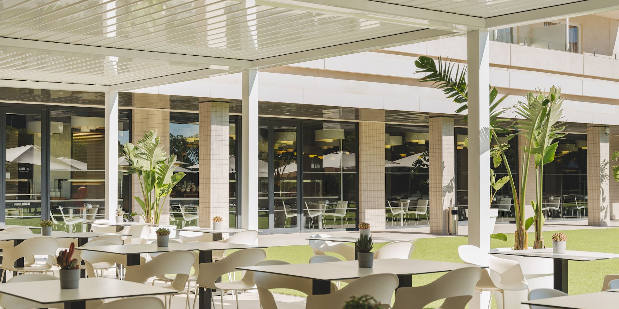 Terraza exterior de restaurante hotelero con mesas, sillas blancas y pérgola moderna.