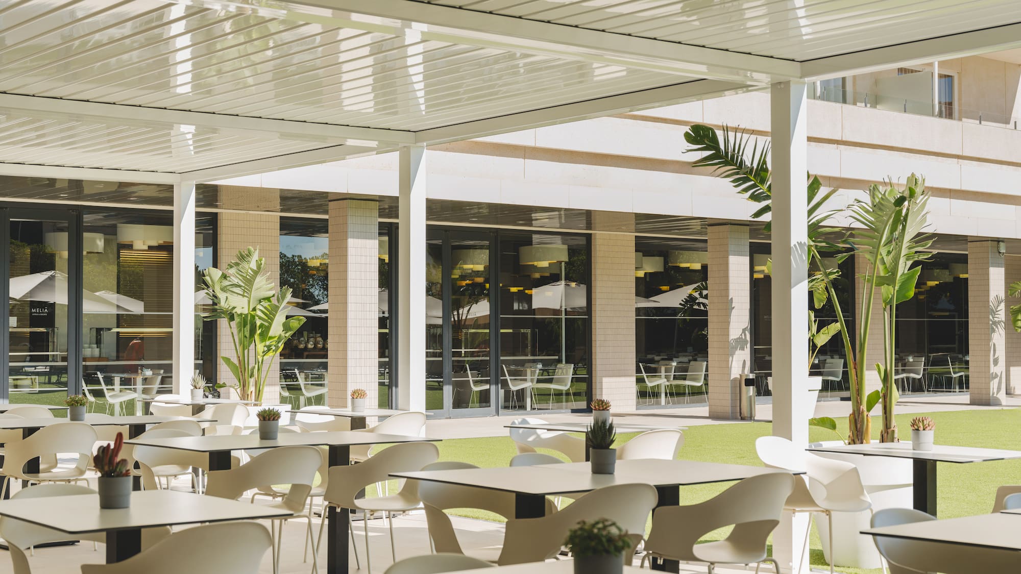 Terraza exterior de restaurante hotelero con mesas, sillas blancas y pérgola moderna.