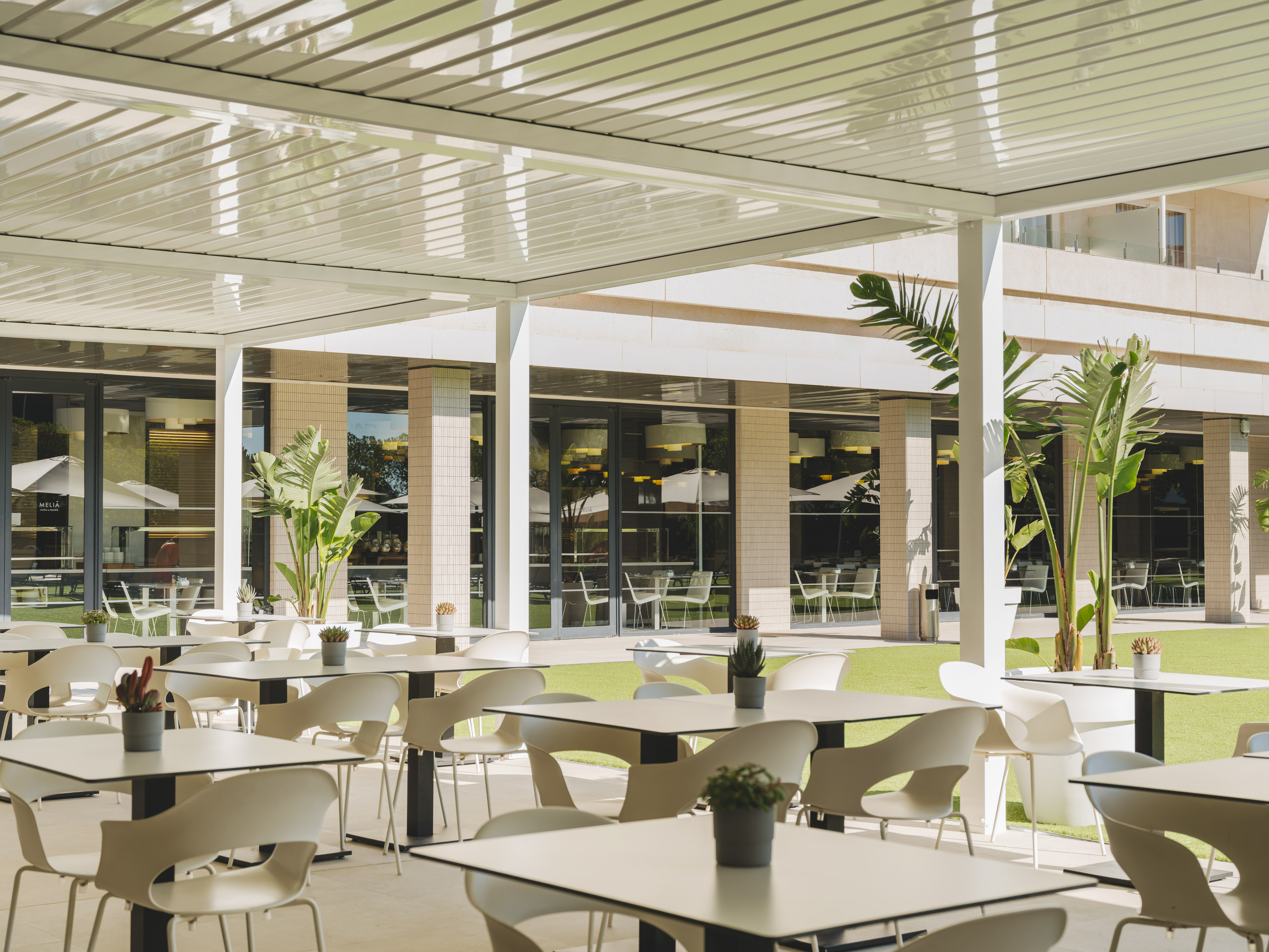 Terraza exterior de restaurante hotelero con mesas, sillas blancas y pérgola moderna.