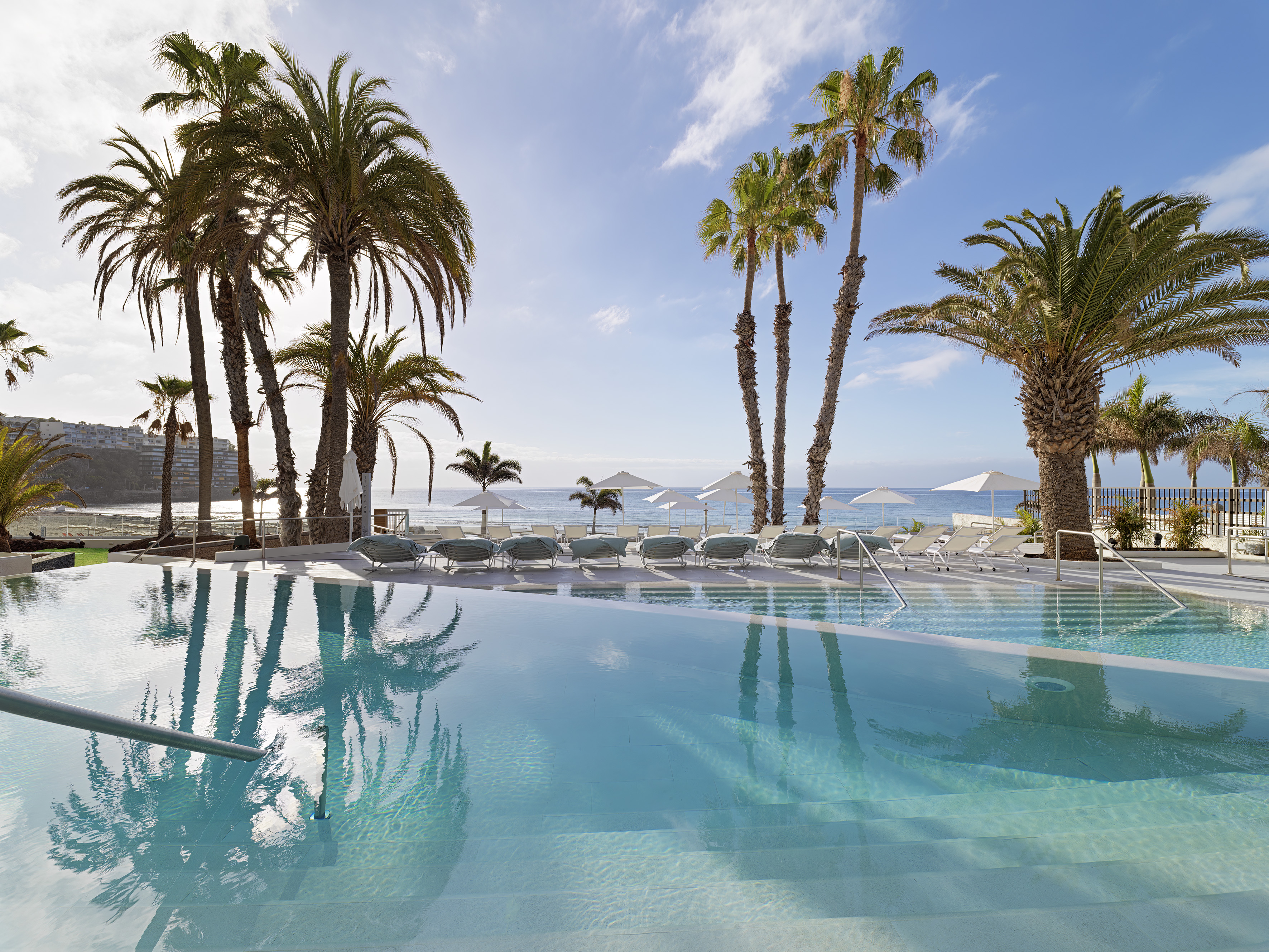 Tropical resort infinity pool with palm trees, ocean, and beach loungers.