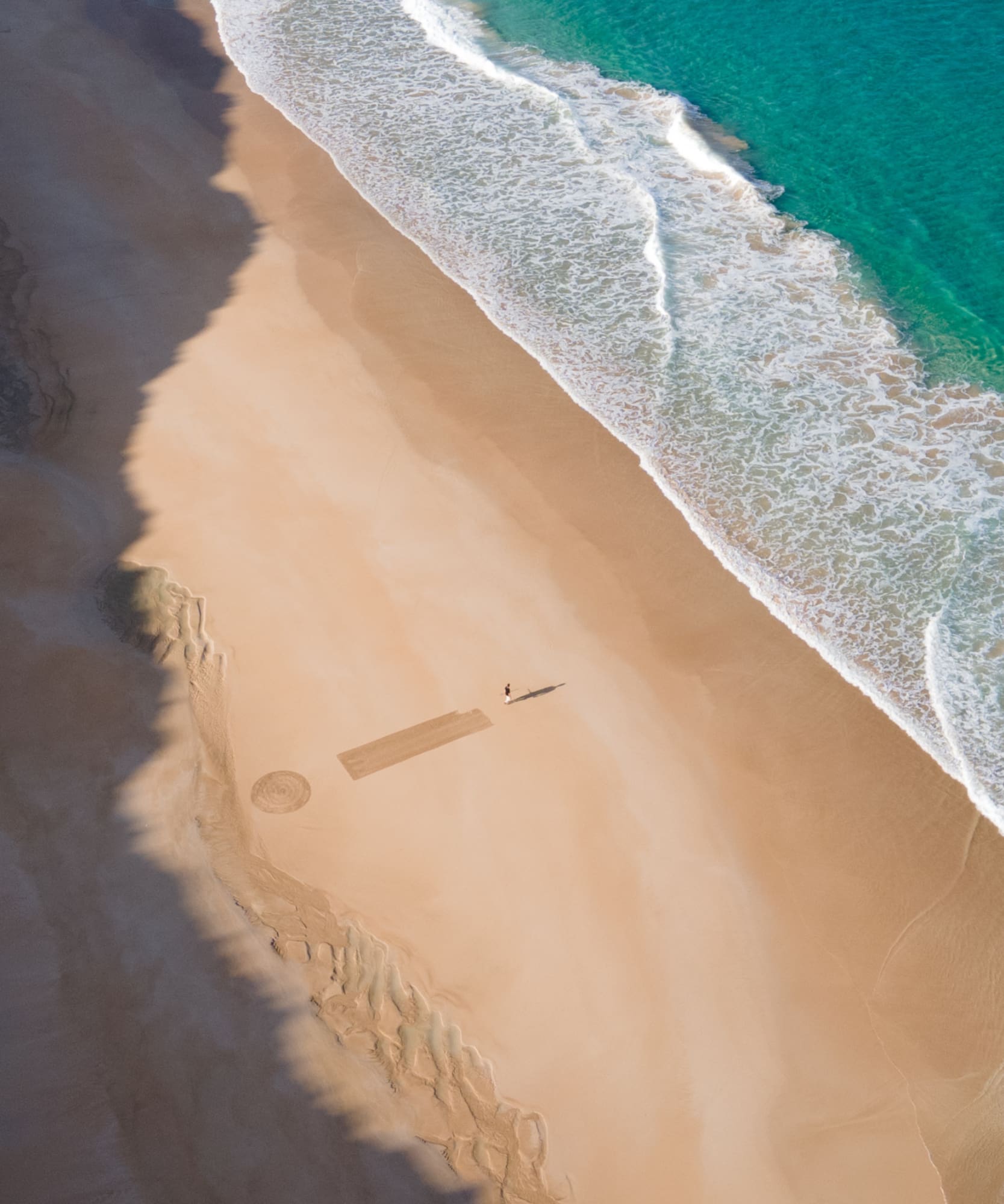 Vista aérea: playa dorada, mar turquesa con olas, persona y arte en arena.