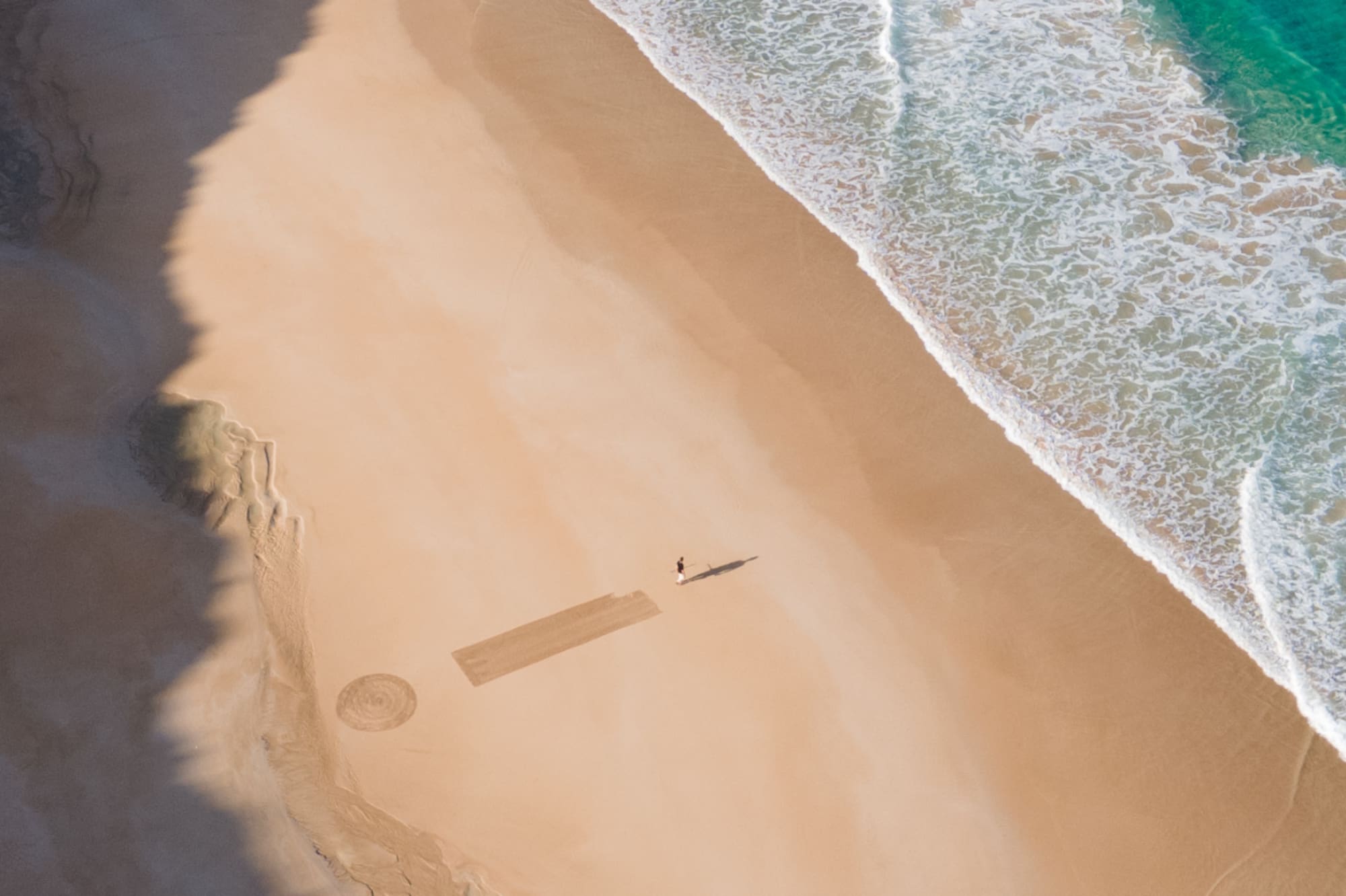 Vista aérea: playa dorada, mar turquesa con olas, persona y arte en arena.