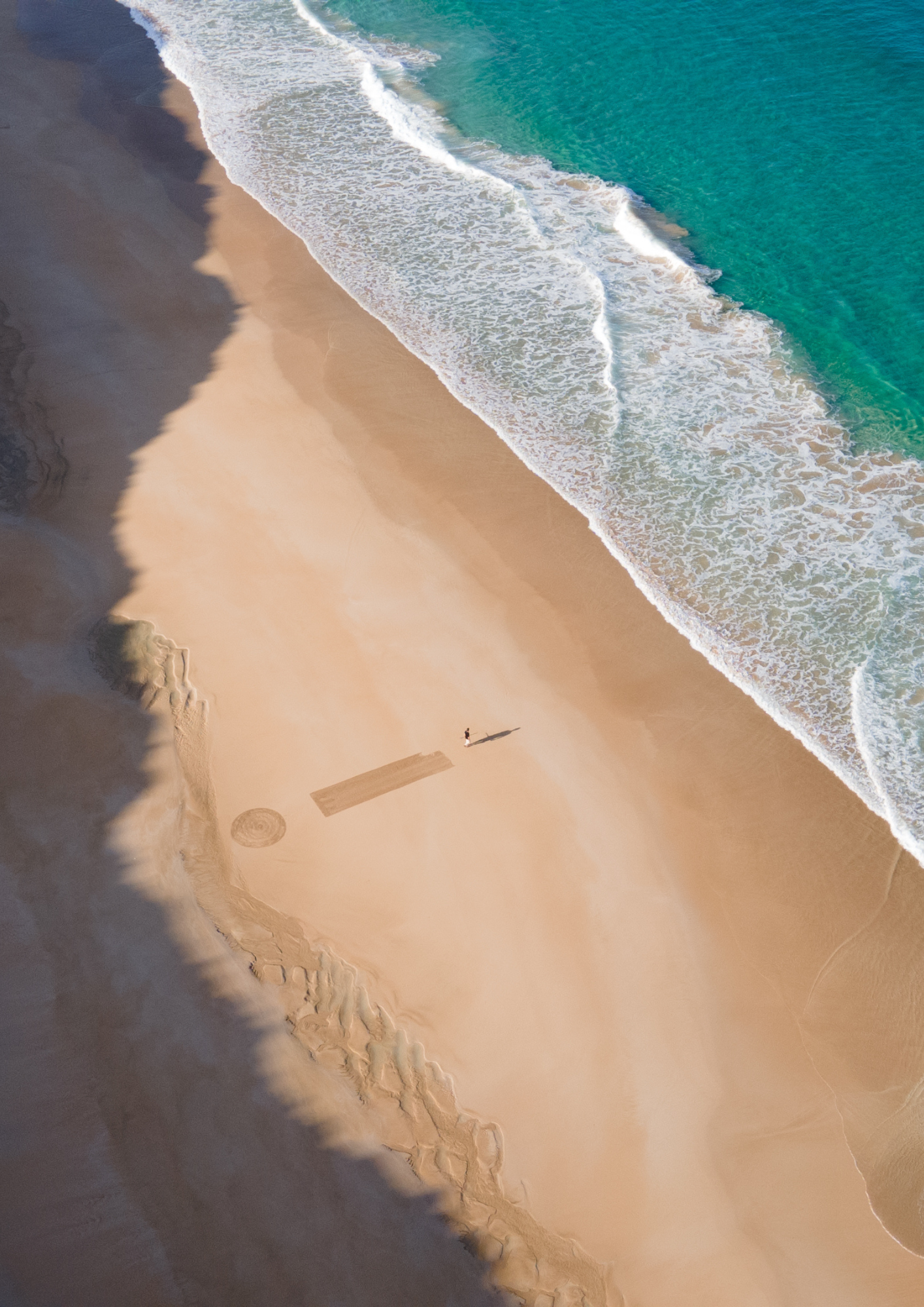 Vista aérea: playa dorada, mar turquesa con olas, persona y arte en arena.