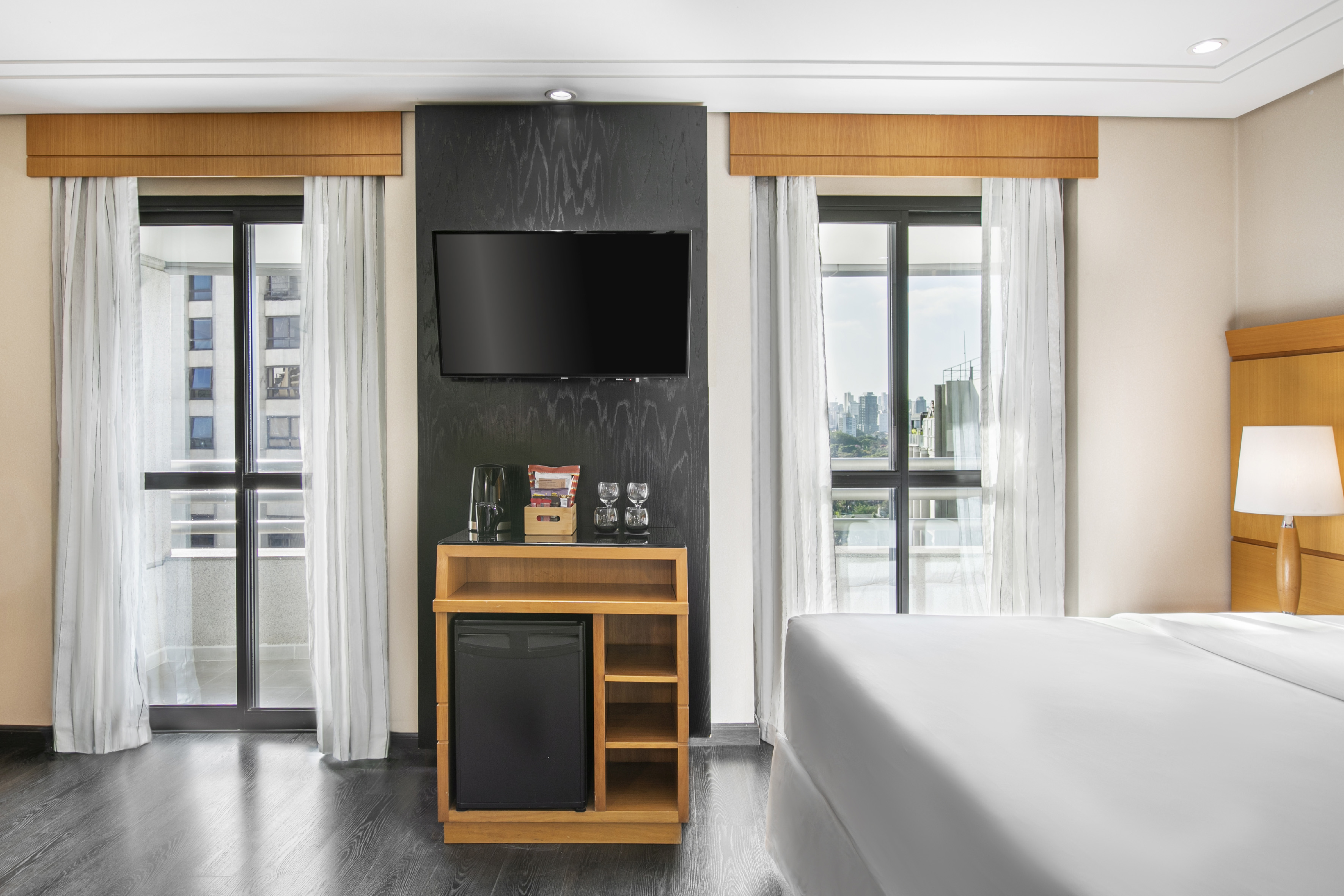Modern hotel bedroom with TV, mini-fridge, and city view from large windows.