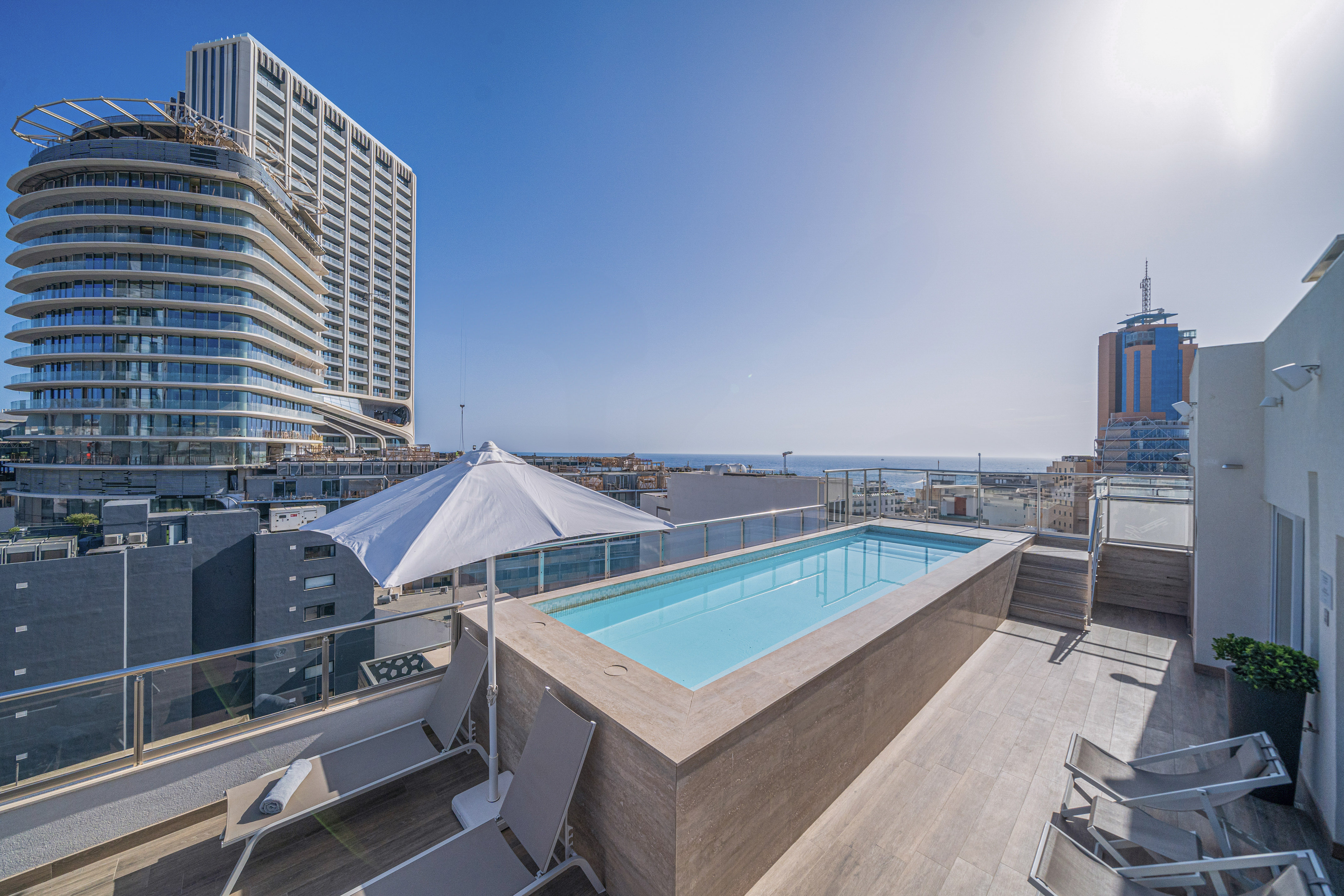 Rooftop plunge pool with lounge chairs, modern city skyline, and ocean view.