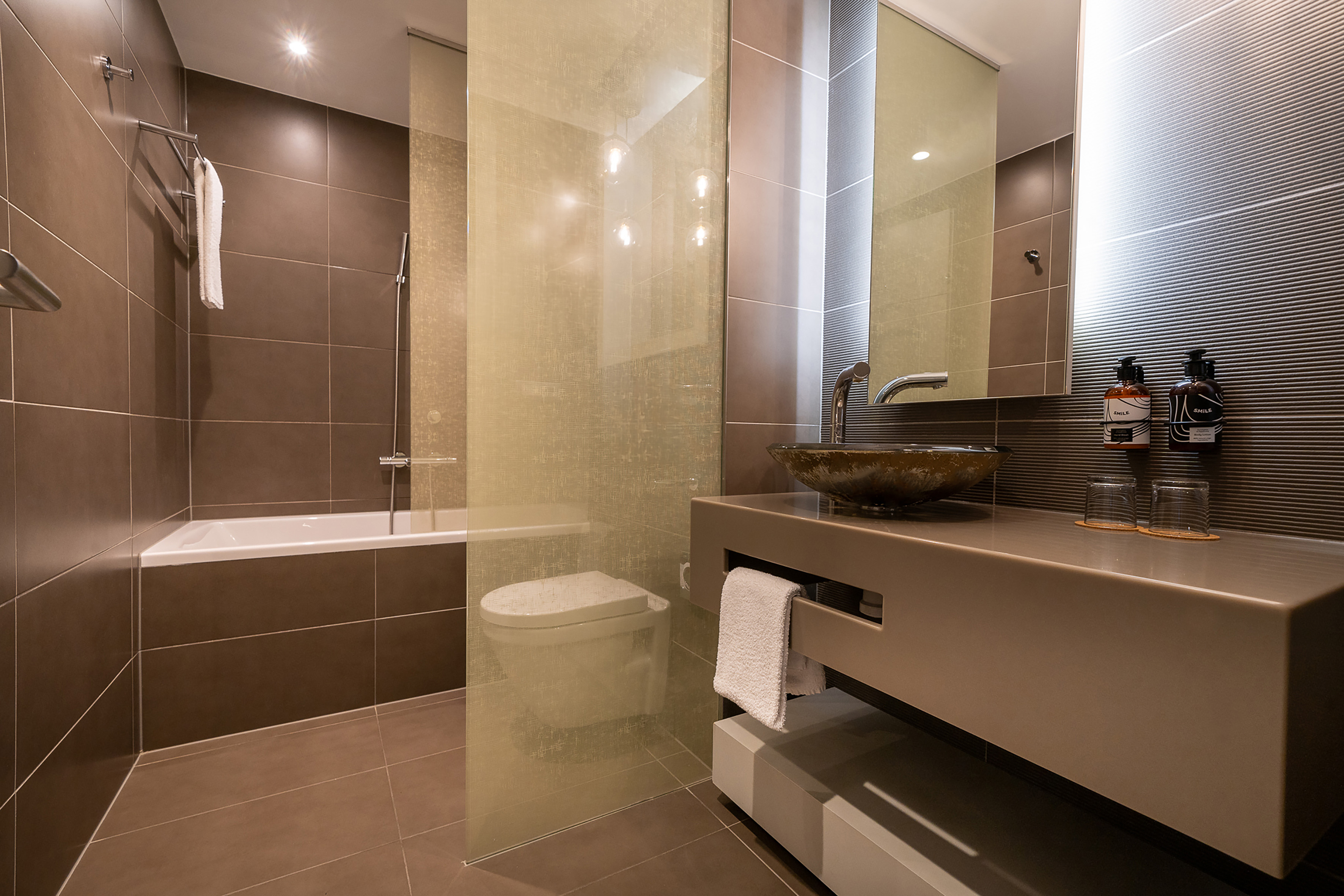 Contemporary bathroom with brown tiled walls, glass basin vanity, and white bathtub.