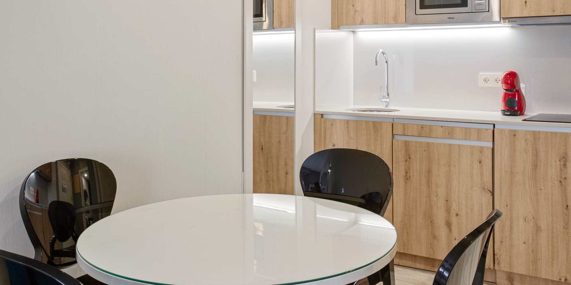 Compact modern kitchen with wood cabinets, white table, and black chairs.