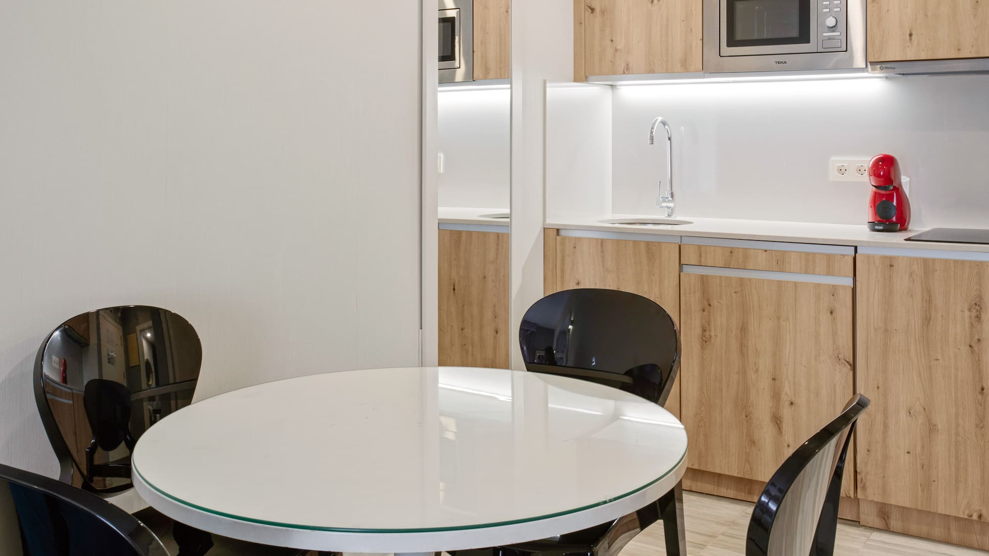 Compact modern kitchen with wood cabinets, white table, and black chairs.