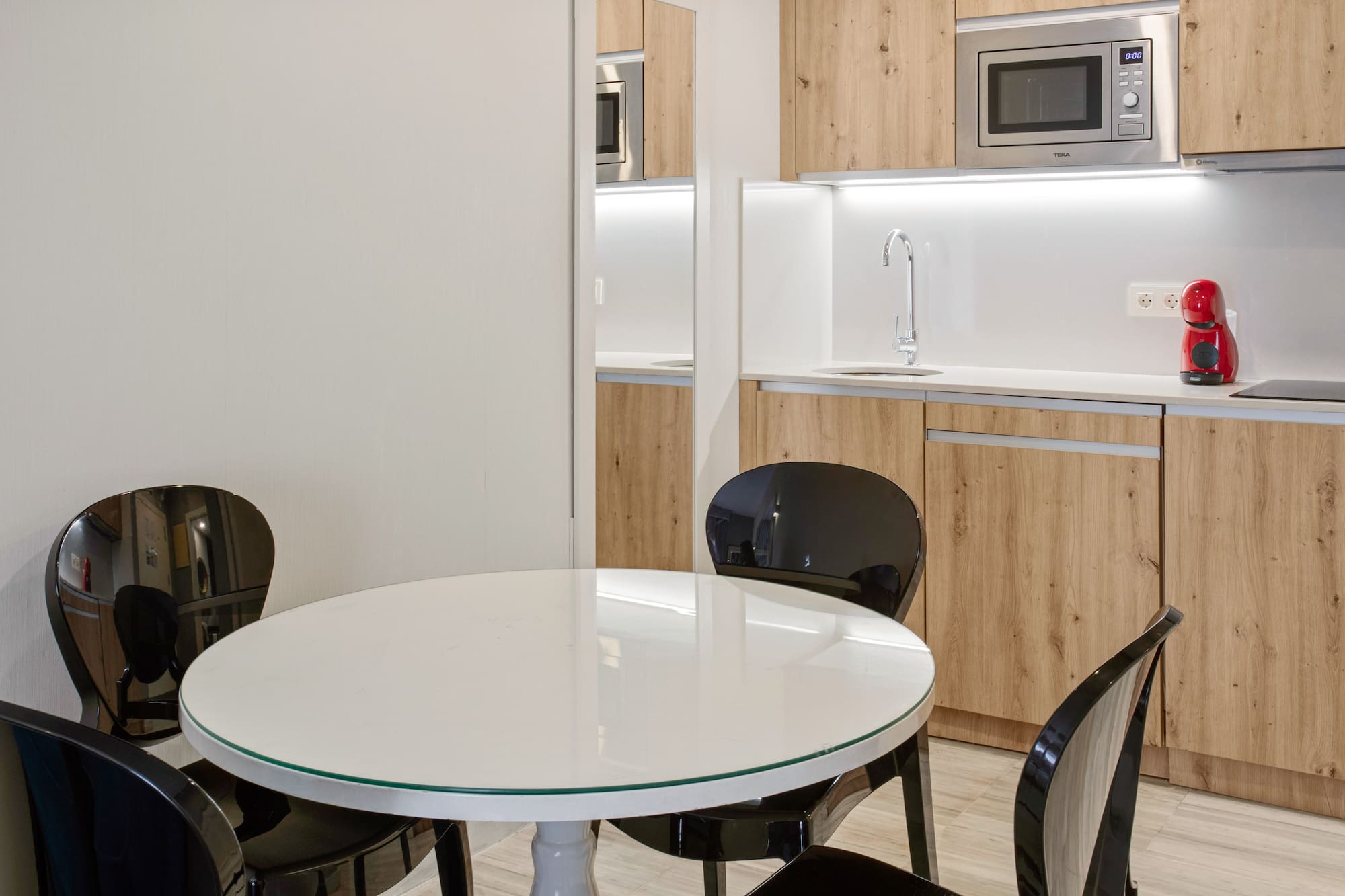 Compact modern kitchen with wood cabinets, white table, and black chairs.