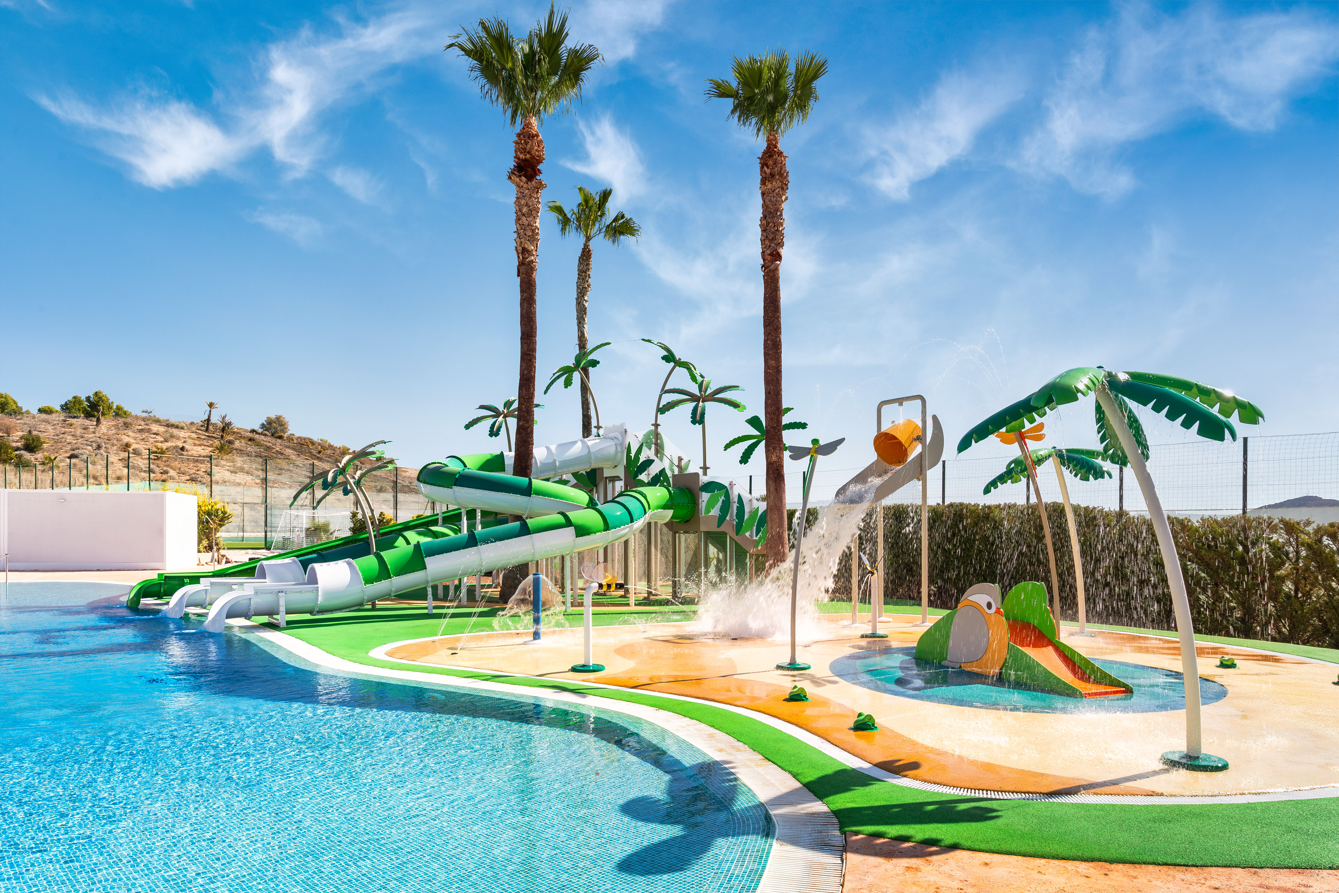Sunny water park with green slides, splash pad, and swimming pool under palm trees.